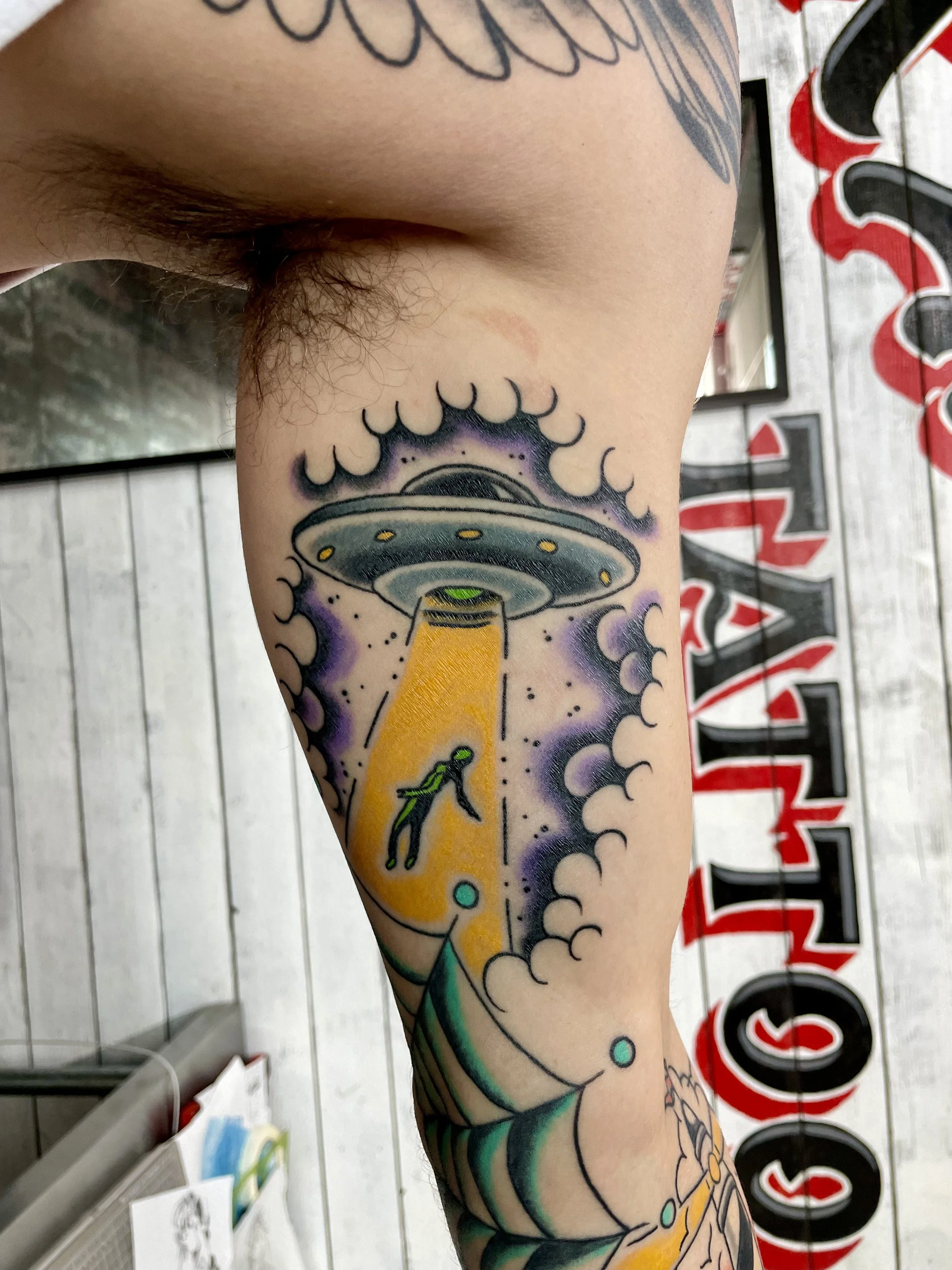 Traditional American color inner bicep tattoo featuring a classic flying saucer with yellow lights beaming down a tractor beam, lifting a small human figure into the air, all surrounded by dramatic purple clouds and swirling smoke, custom tattoo by N