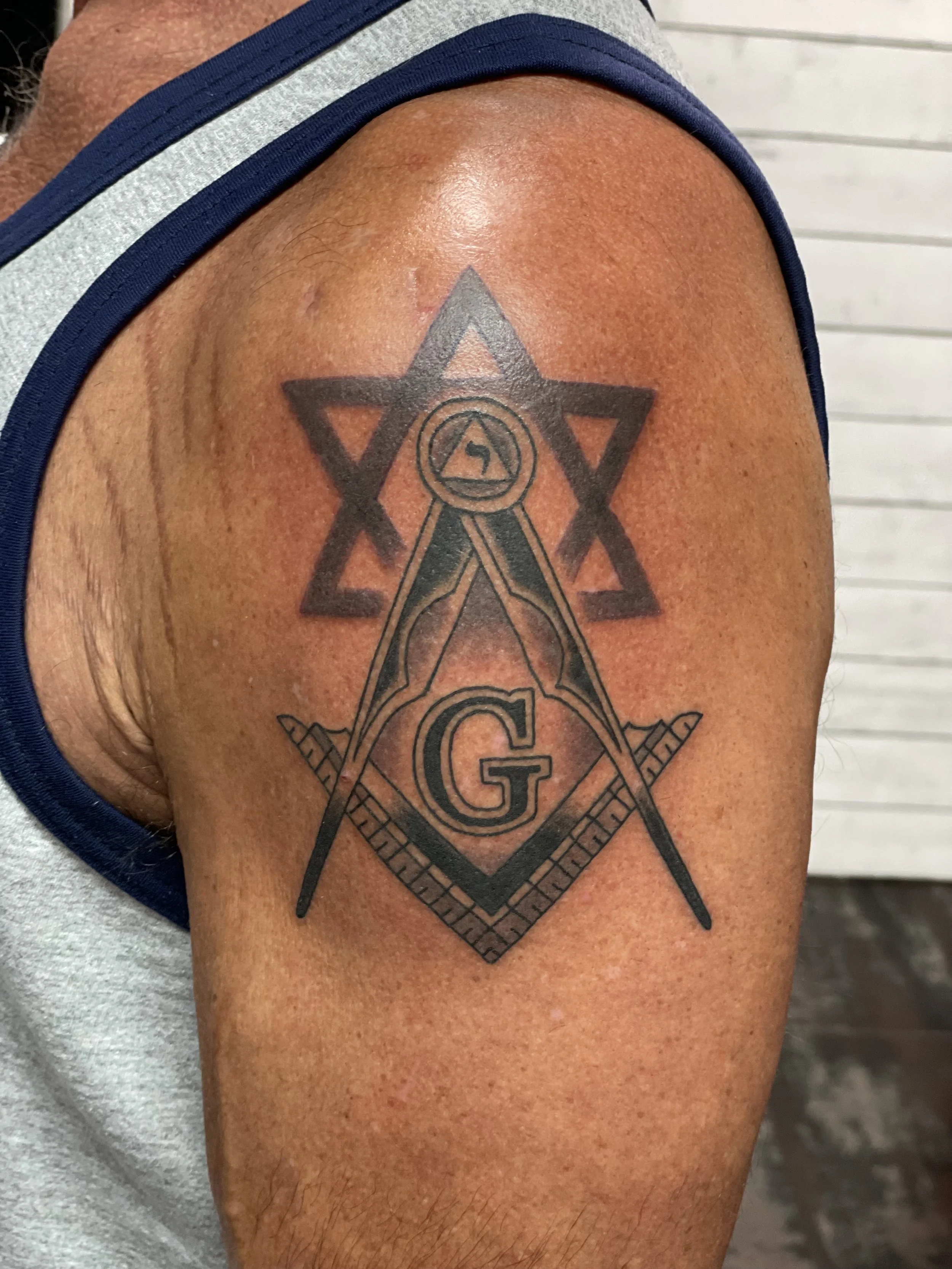 Two of the most recognizable symbols in fraternal history layered into one powerful shoulder piece. The Masonic square and compass centered over a bold Star of David, with a compass and triangle at the apex. Black and grey shoulder tattoo by Nick at 