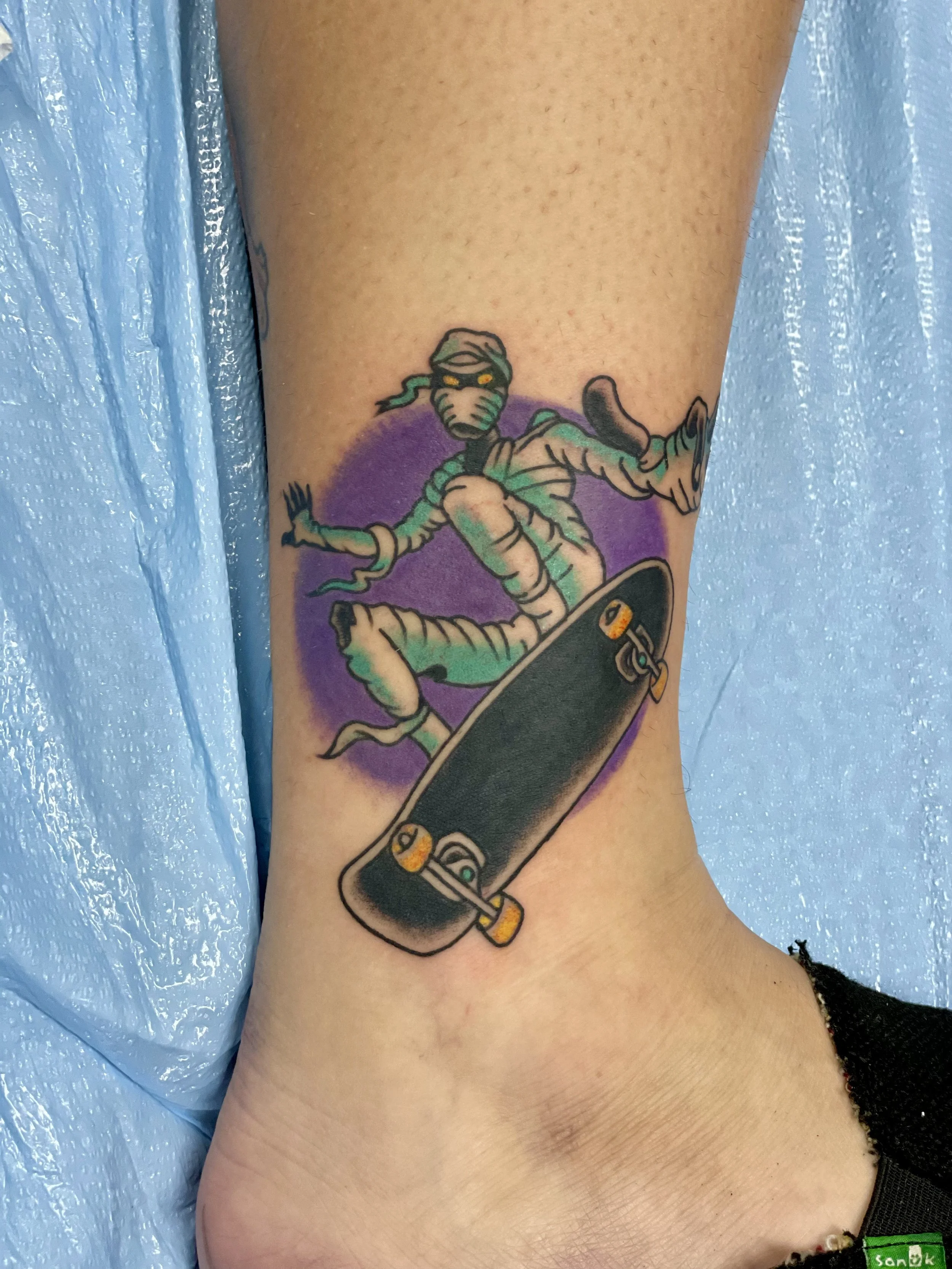 Traditional American color ankle tattoo featuring a wrapped mummy with glowing yellow eyes performing a skateboard trick, set against a bold purple circle background, custom tattoo by Nick at Big Kahuna Tattoo in Boca Raton, FL.