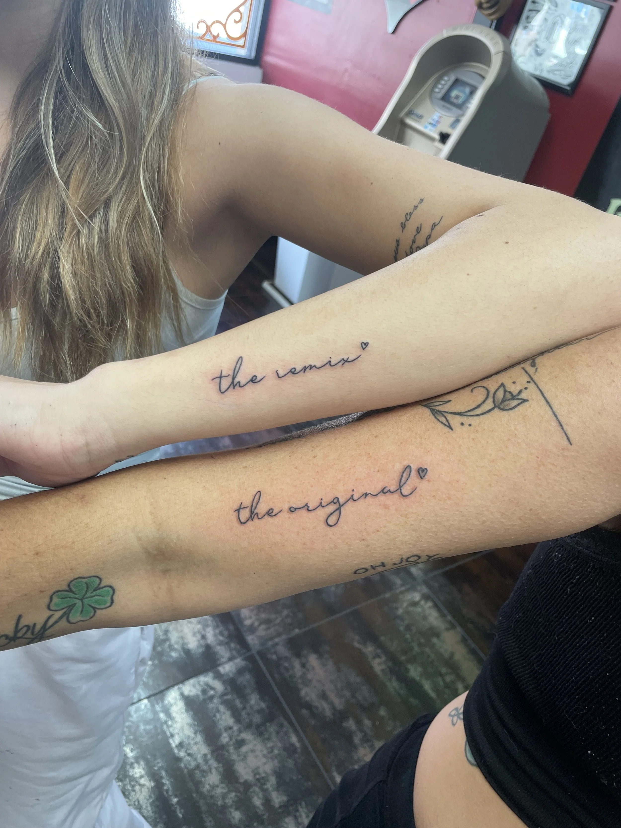 Matching fine line forearm tattoos for two, featuring delicate cursive script reading "the original" and "the remix," each accented with a small heart, custom matching tattoos by Nick at Big Kahuna Tattoo in Boca Raton, FL.