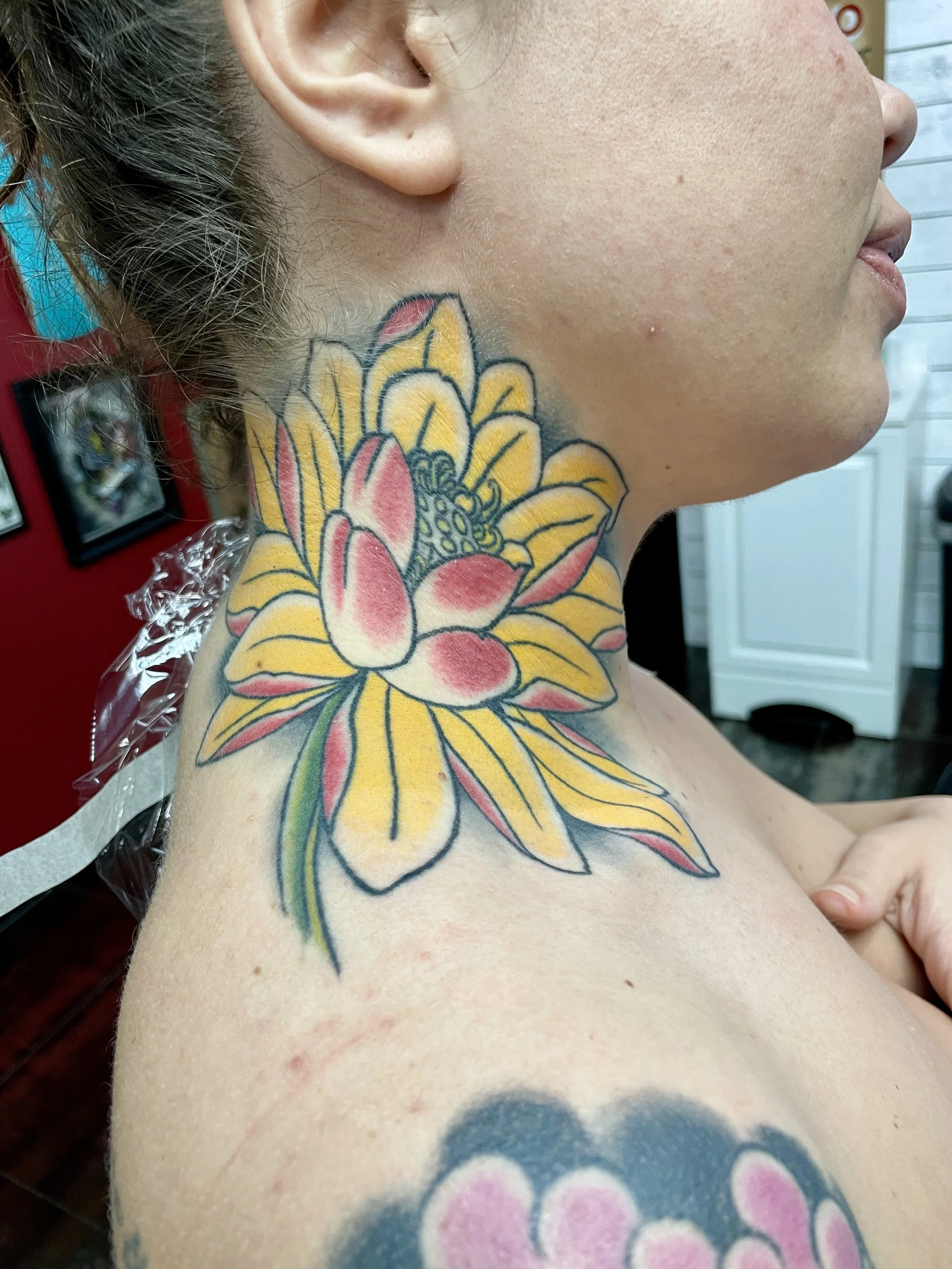 Large traditional American color neck tattoo featuring a blooming lotus flower with vibrant yellow inner petals and soft red outer petals surrounding a detailed seed pod center, custom tattoo by Nick at Big Kahuna Tattoo in Boca Raton, FL.
