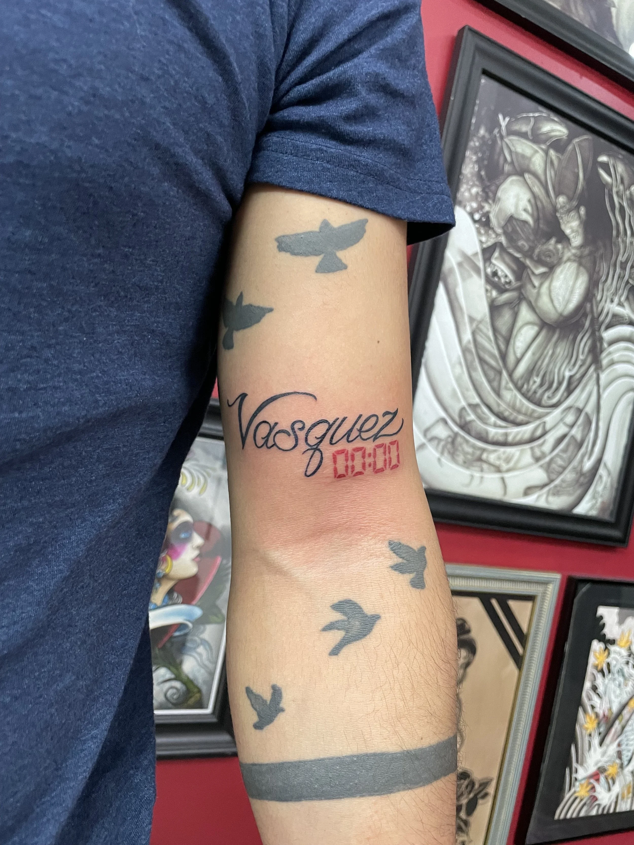 Fine line forearm tattoo featuring the script name "Vasquez" with a red digital clock reading "00:00" beneath it, surrounded by silhouette birds in flight scattered above and below, with a solid black band at the wrist, custom tattoo by Nick at Big K