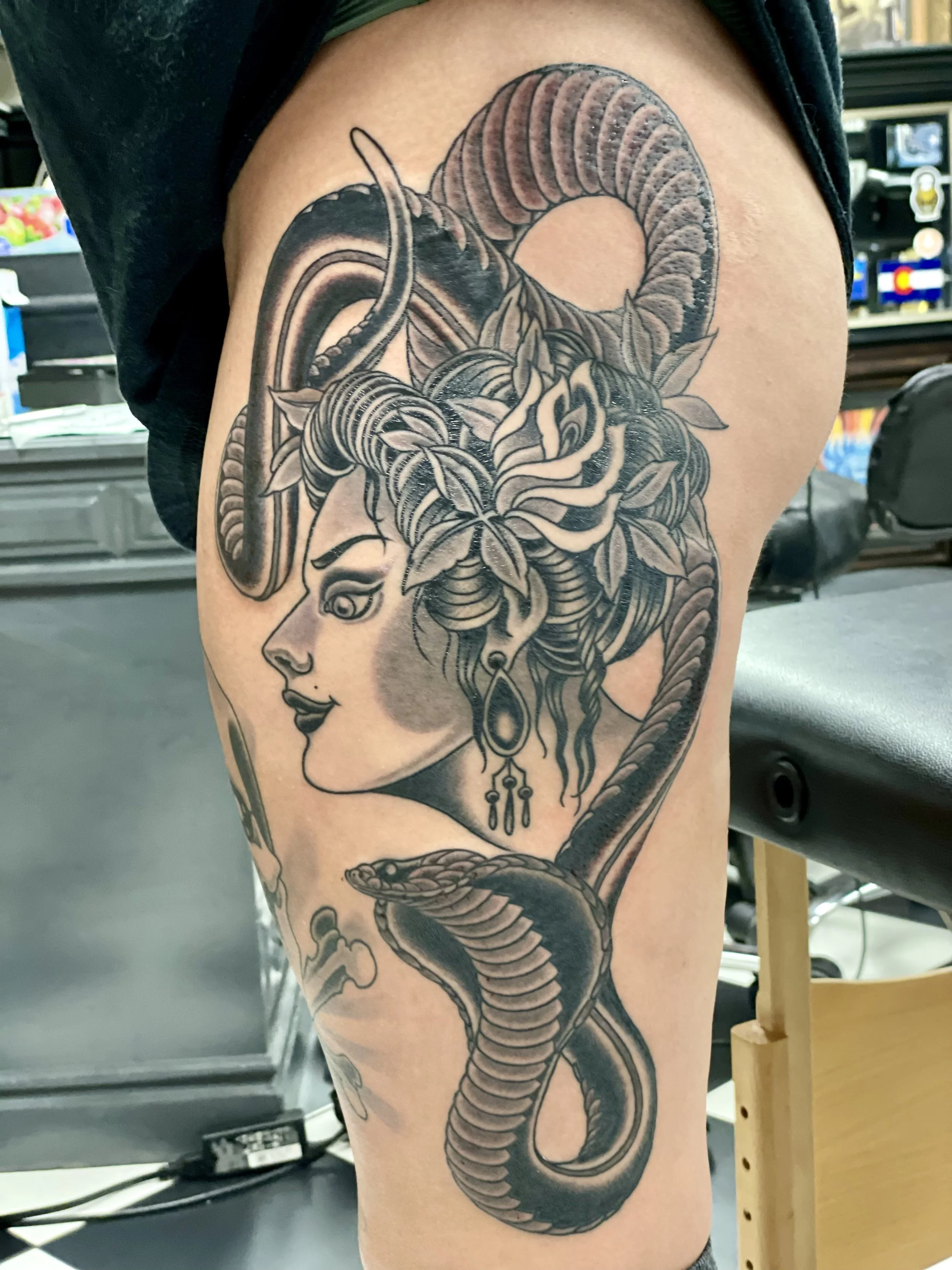 A commanding thigh piece featuring a goddess in a side profile with flowers and leaves woven through her hair, jeweled earrings, and a coiling cobra rising below her. dark, detailed, and undeniably striking. Black and grey thigh tattoo by Nick at Big
