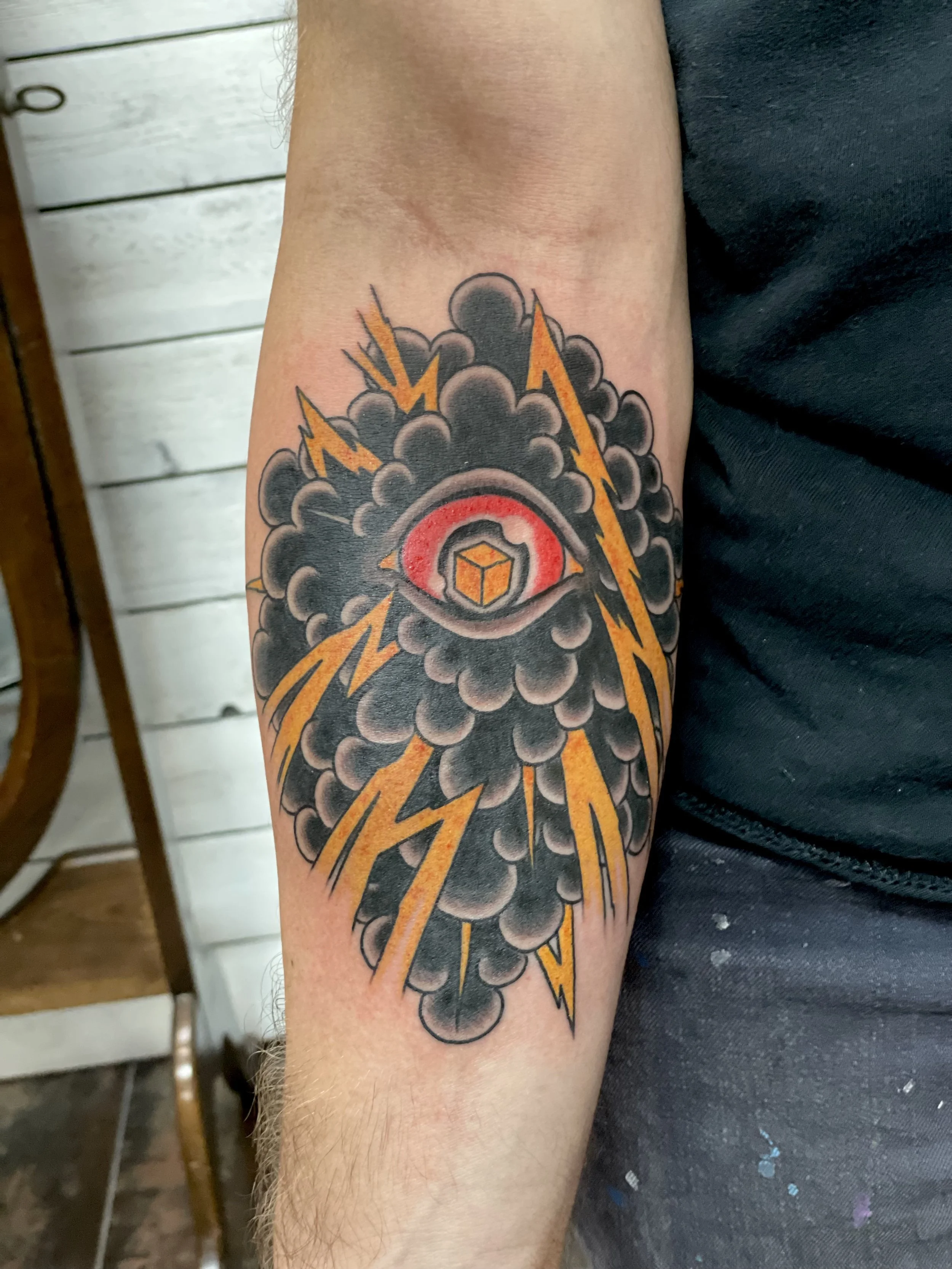 Traditional American color forearm tattoo featuring an all-seeing eye with a red iris and geometric cube pupil, centered within a dark storm cloud mass crackling with bold gold lightning bolts, custom tattoo by Nick at Big Kahuna Tattoo in Boca Raton
