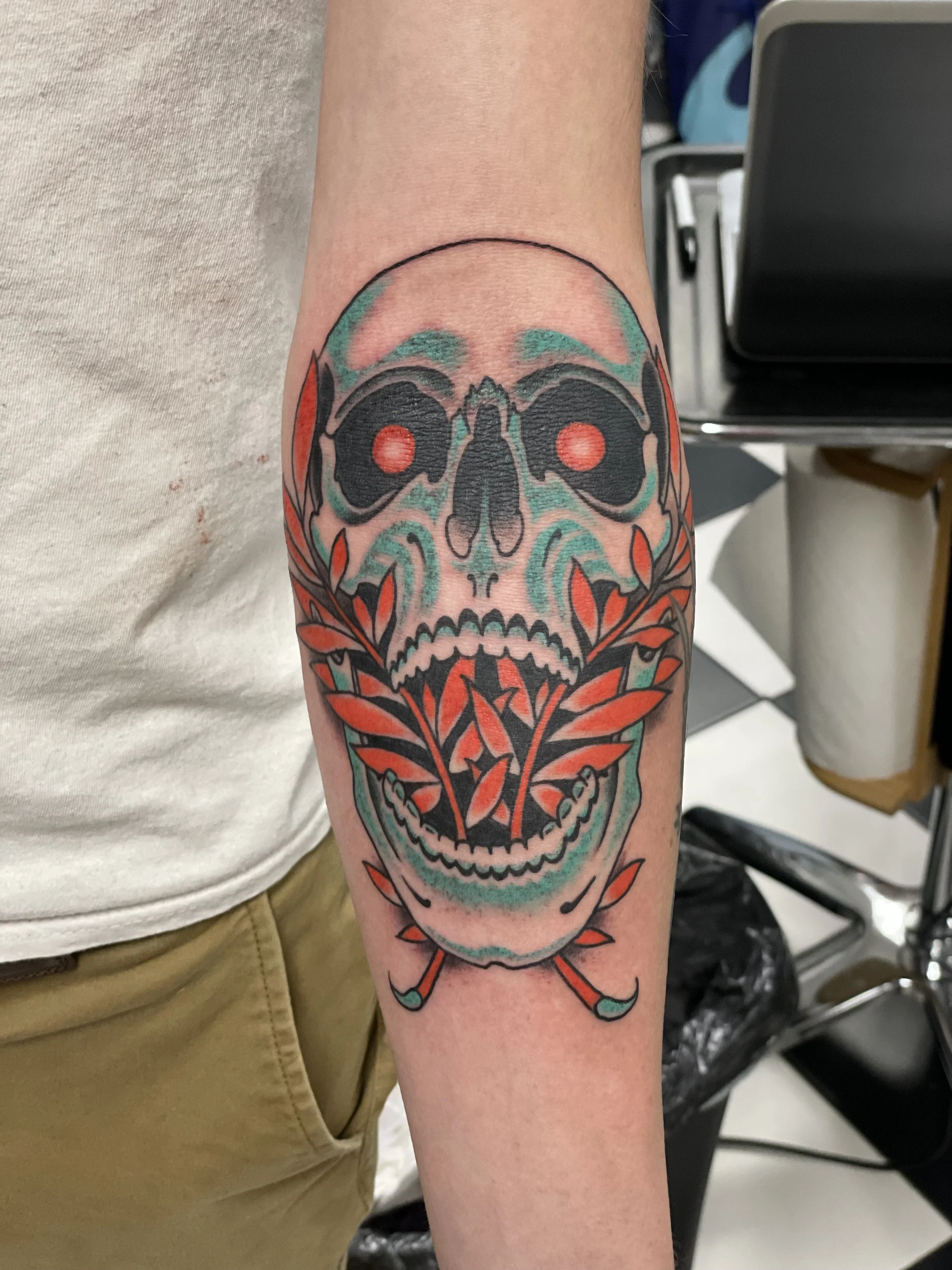 Traditional American color tattoo featuring a large teal skull with glowing red eyes, framed by bold red and orange laurel branches and crossed bones below, custom tattoo by Nick at Big Kahuna Tattoo in Boca Raton, FL.
