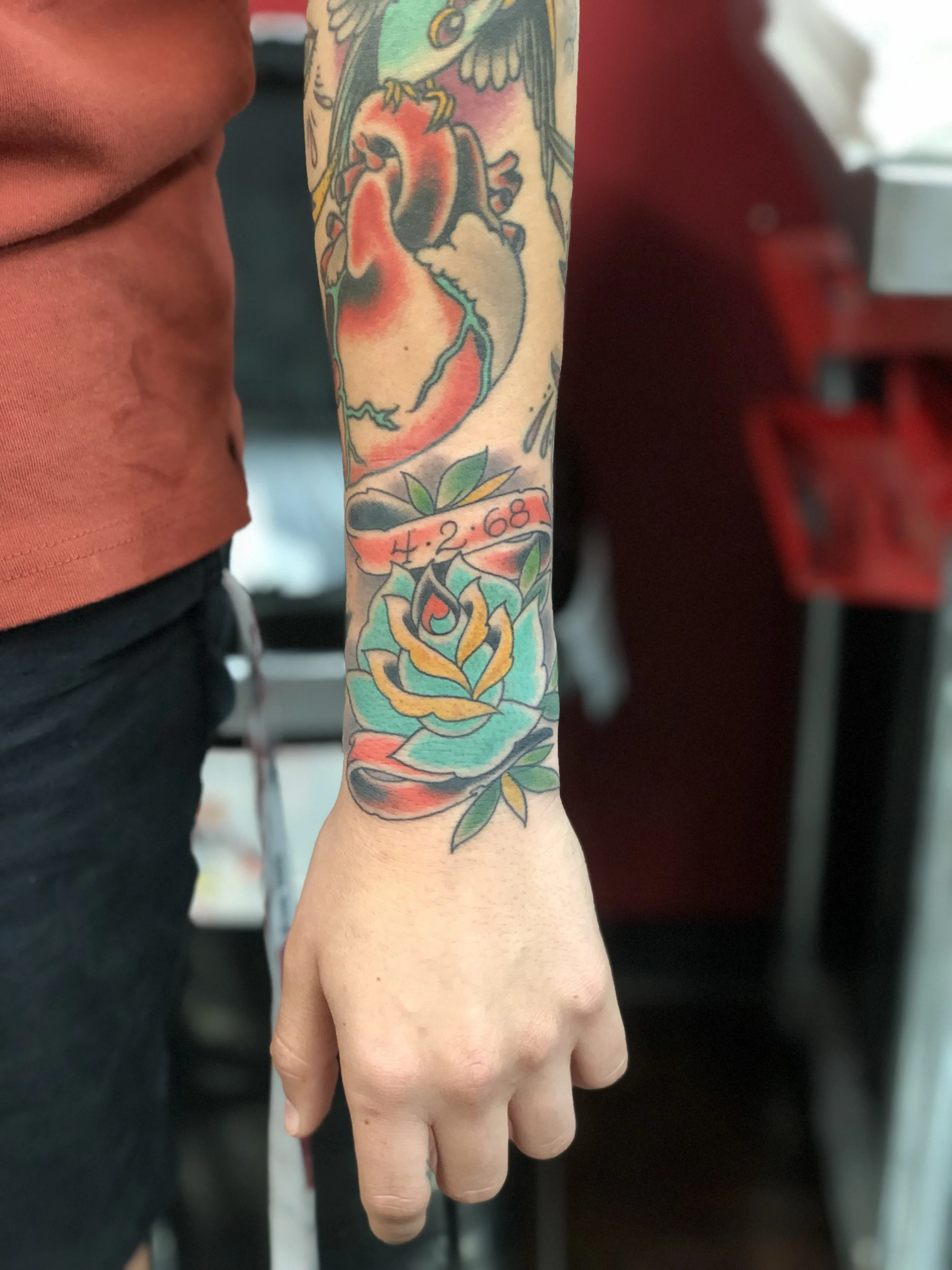 Traditional American color tattoo featuring a teal rose with a red banner reading "4.2.68" on the wrist, part of a larger sleeve, custom tattoo by Nick at Big Kahuna Tattoo in Boca Raton, FL.