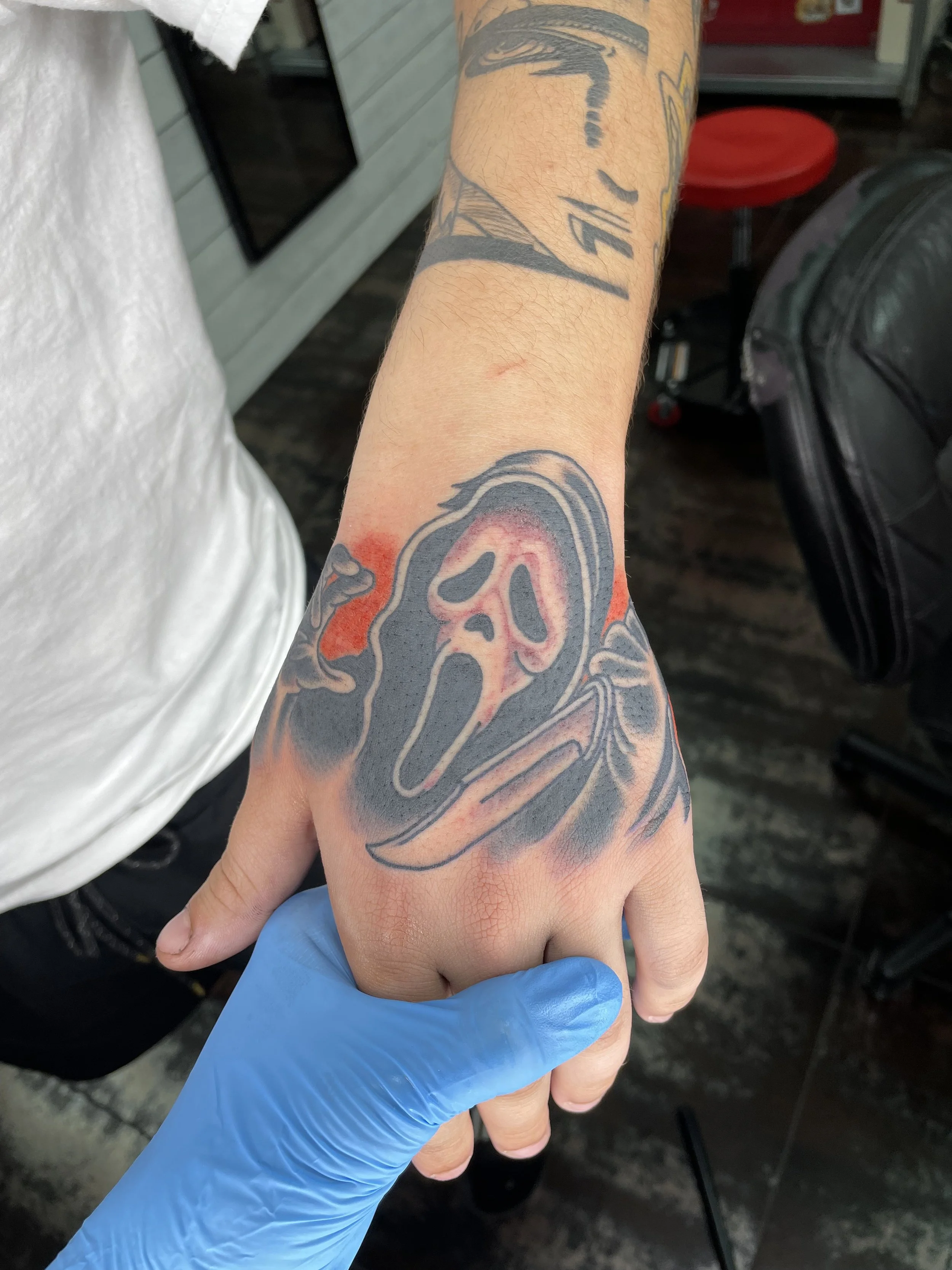 Black and grey hand tattoo featuring the iconic Ghostface mask from the Scream franchise, rendered in bold linework with a red accent halo behind the mask and a knife in hand, custom tattoo by Nick at Big Kahuna Tattoo in Boca Raton, FL.