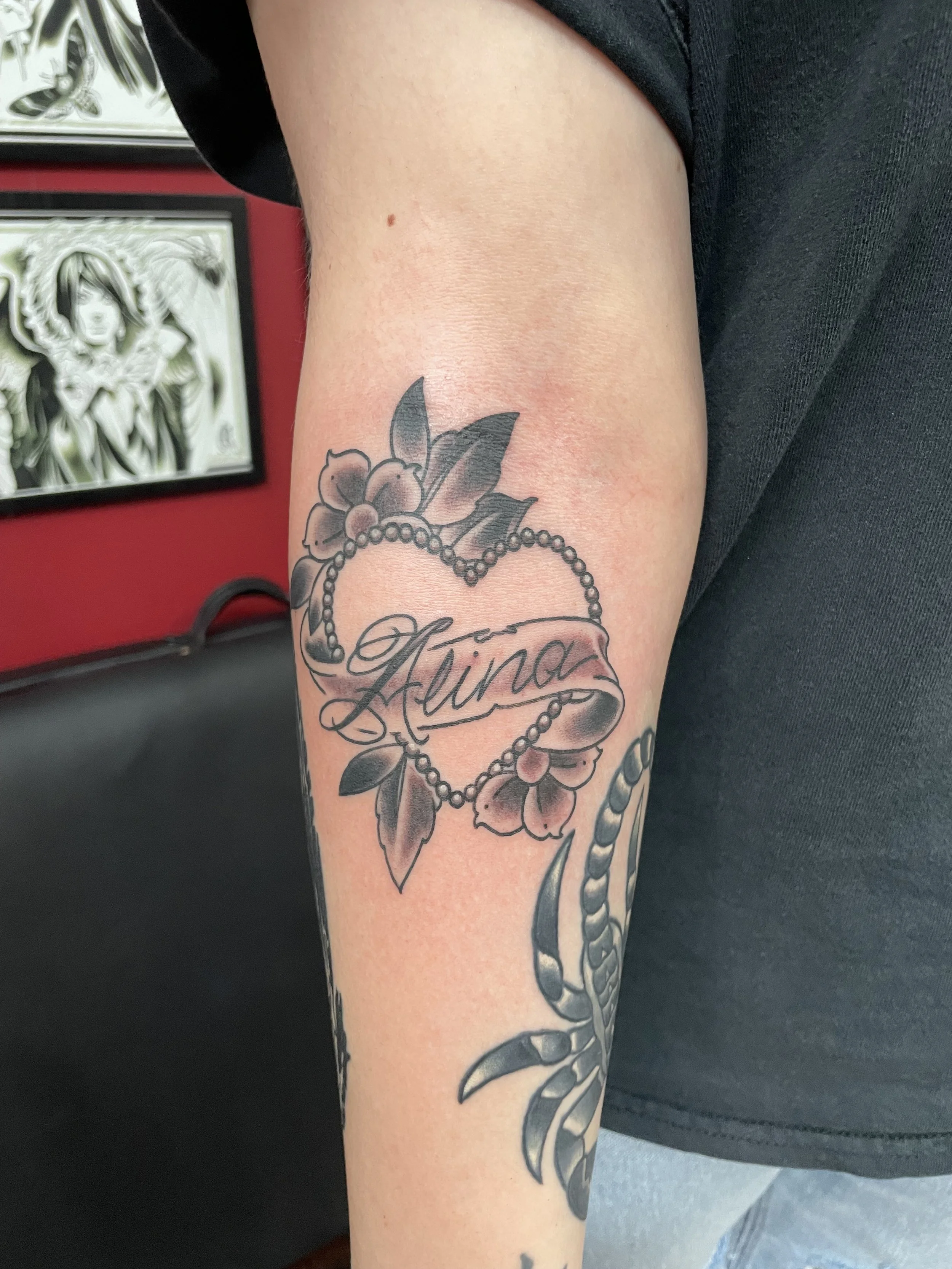 A black and grey forearm piece built around a pearl beaded heart outlined in small blossoms and leaves, with the name "Alina" scripted across a flowing banner through the center. Personal, feminine, and beautifully composed. Forearm tattoo by Nick at