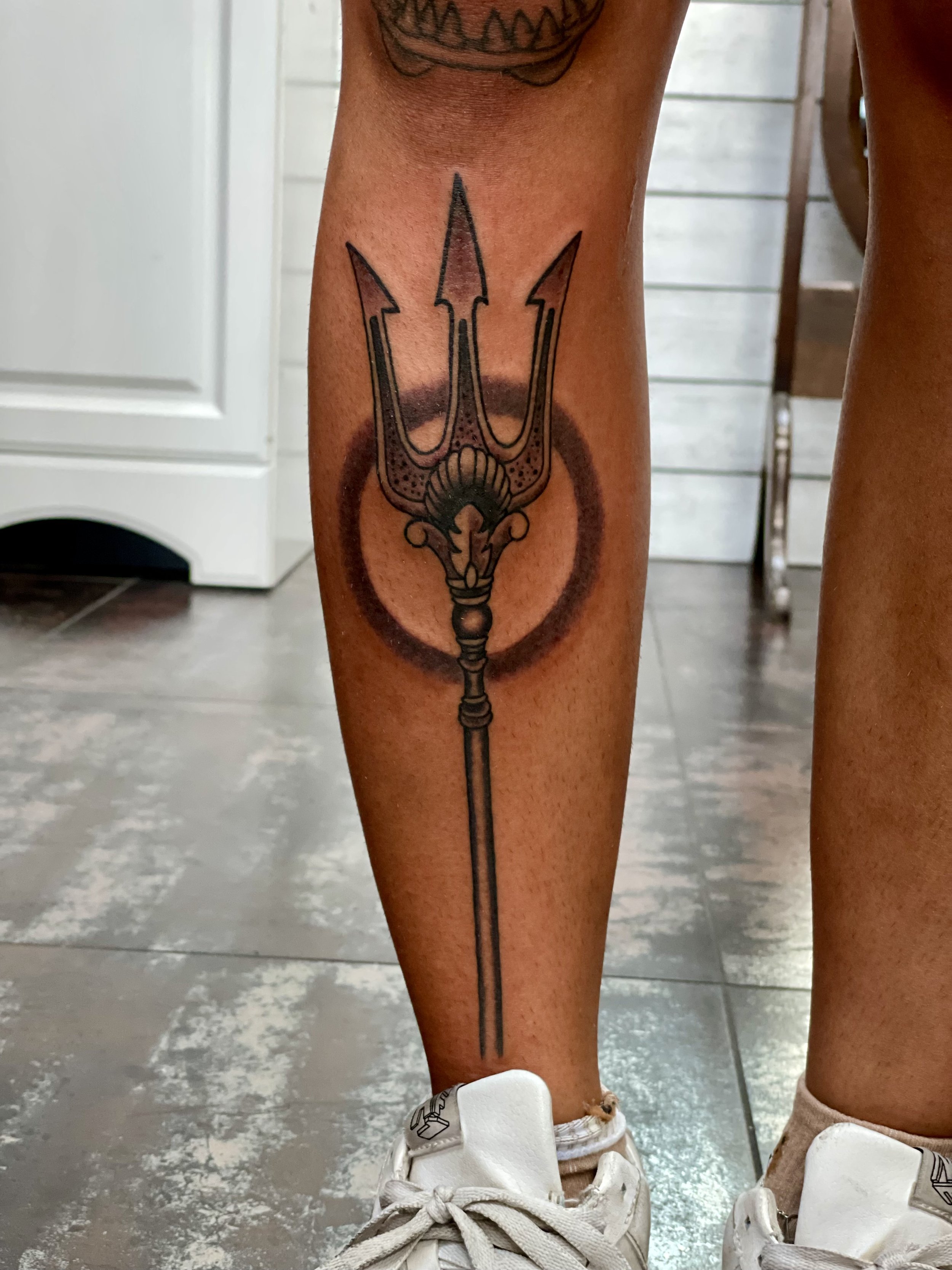 Poseidon's weapon rendered with the precision of an engraving. An ornate trident with a decorative shell crossguard centered in a bold circular shadow halo, the handle running the full length of the shin. Black and grey shin tattoo by Nick at Big Kah