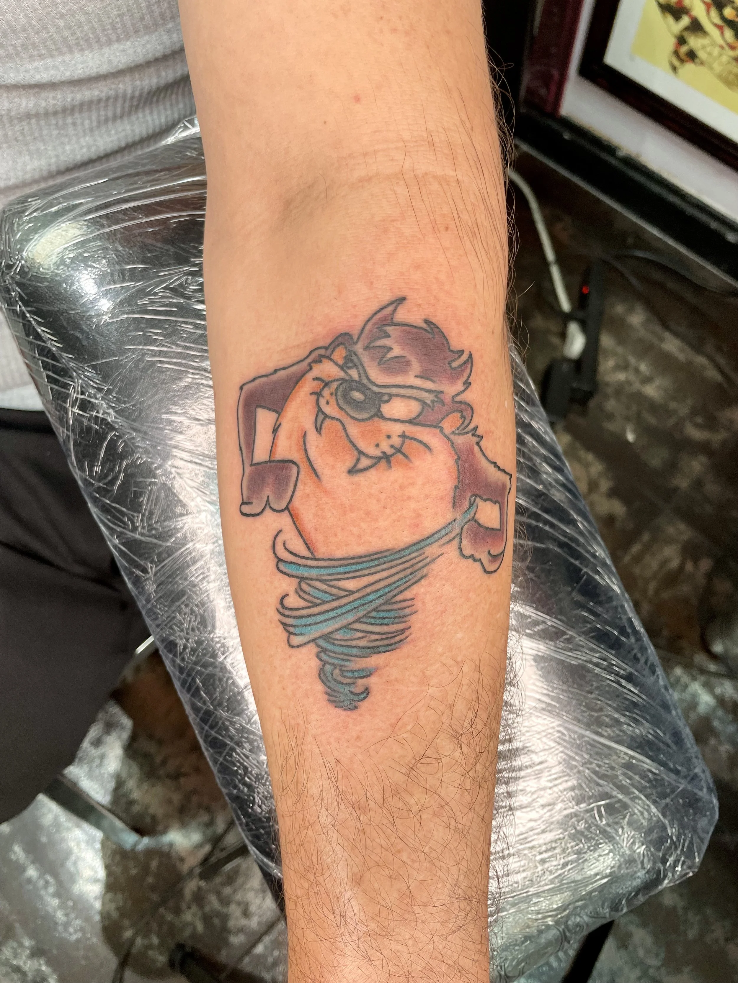 Color cartoon forearm tattoo featuring the Tasmanian Devil spinning in his signature whirlwind tornado, rendered in warm oranges, brown, and teal, custom tattoo by Nick at Big Kahuna Tattoo in Boca Raton, FL.