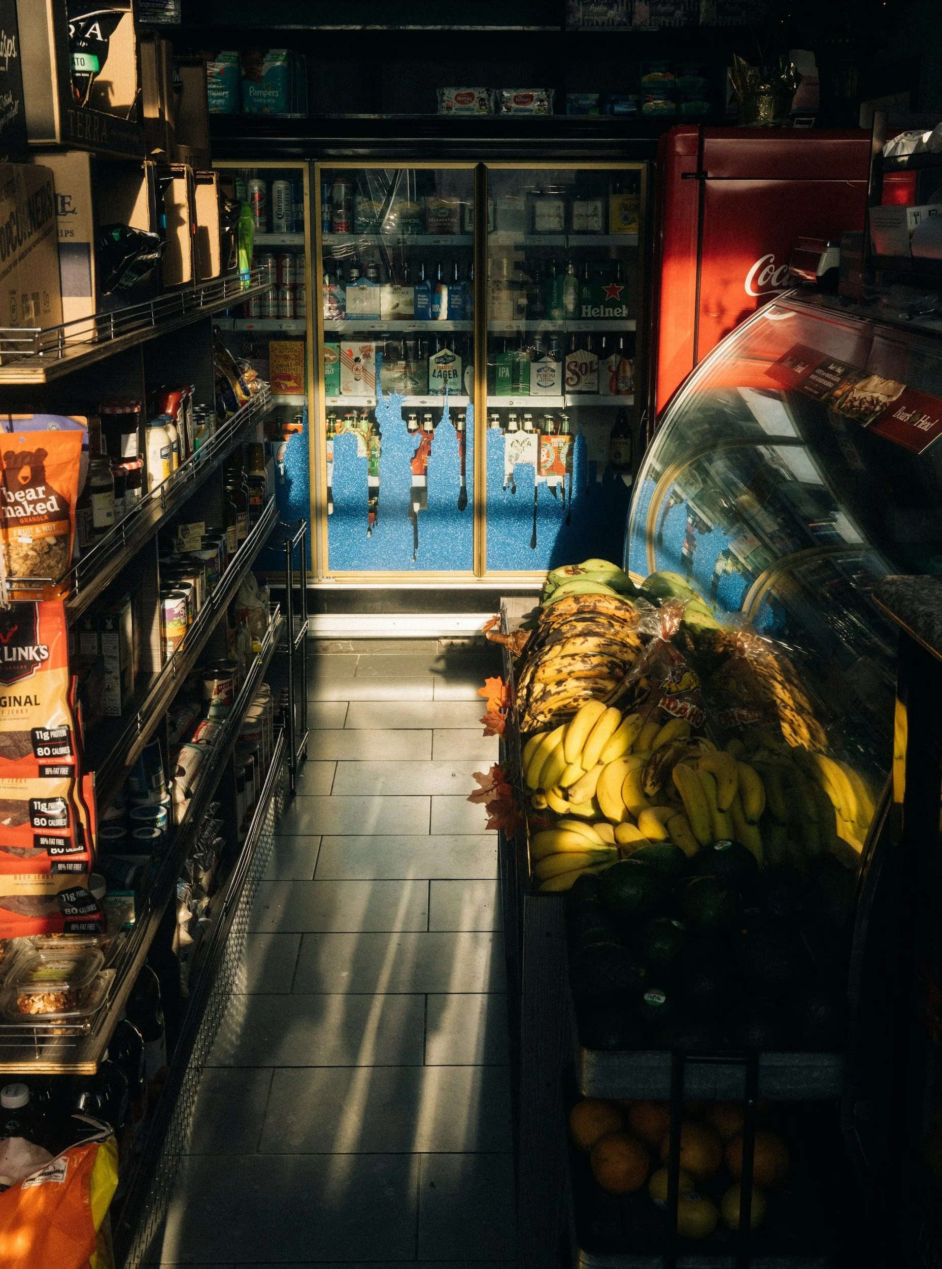 Inside a convenience store, showing shelves with snacks and beverages on the left, a display of bananas and avocados on the right, and a glass door refrigerator in the back containing bottles and cans, with a red Coca-Cola branded cooler next to it.