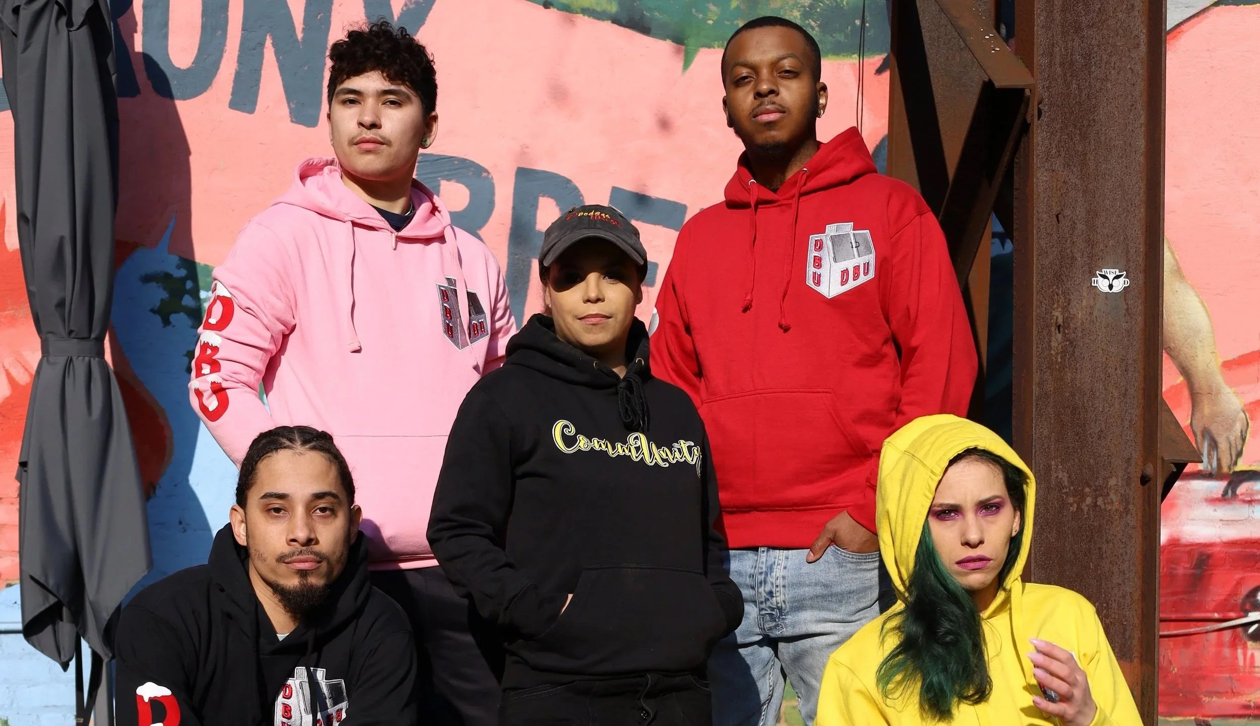 Group of five young adults standing outdoors in front of a colorful mural, wearing hoodies and casual clothing.