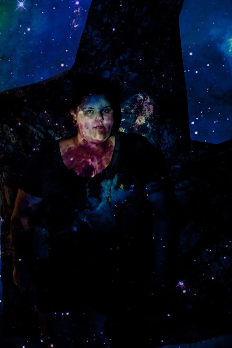 A woman with short dark hair and earrings, sitting against a background with galaxy and stars projections, creating a cosmic visual effect.