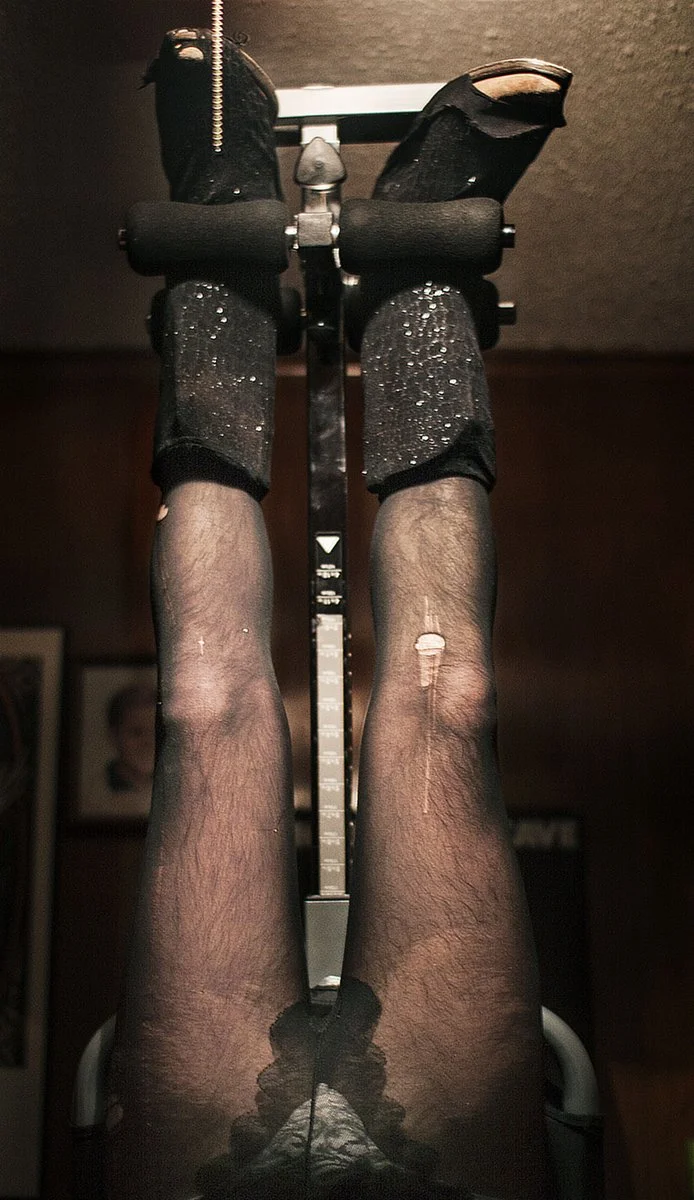 A person with hairy legs wearing torn black shorts and black fishnet stockings, suspended by there feet on an inversion table.