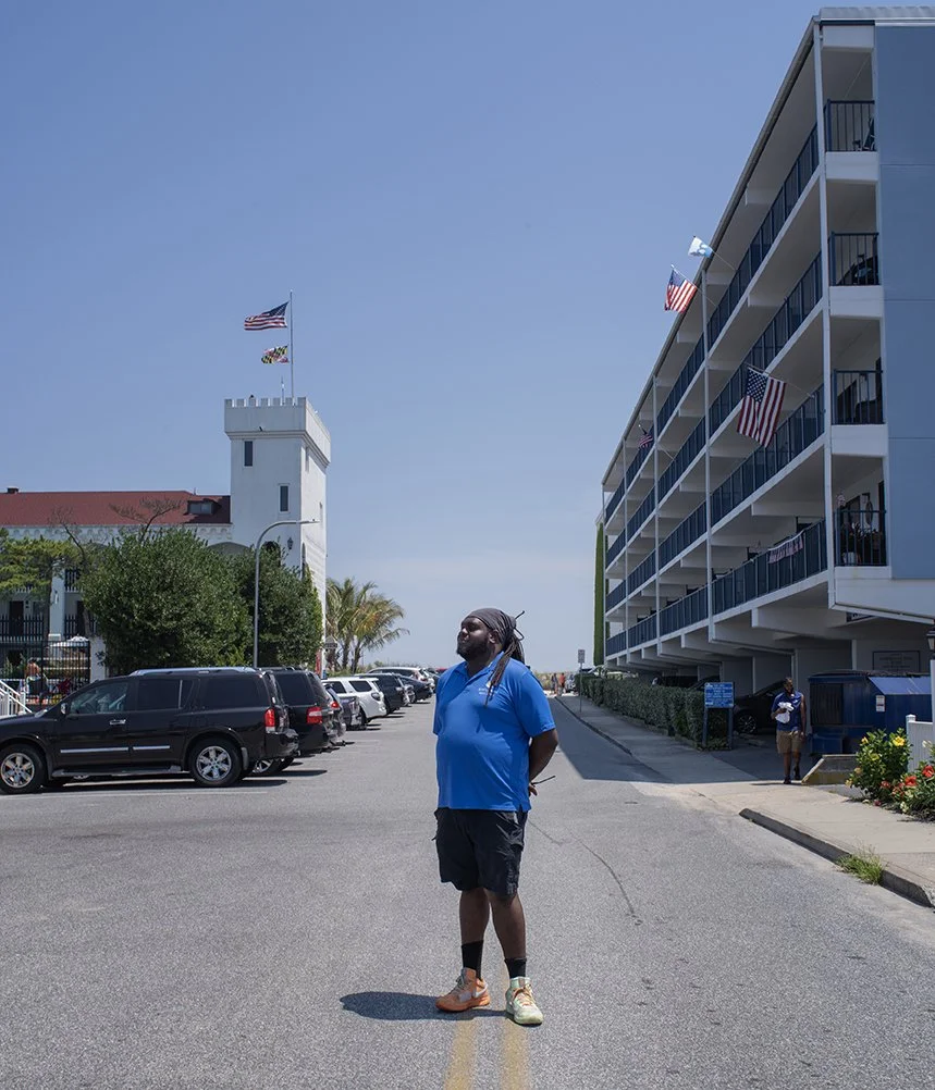 A man with long braids, wearing a blue t-shirt, black shorts, and sneakers, standing in the middle of a parking lot under a clear blue sky, with an apartment building and a historic-looking tower with flags in the background.