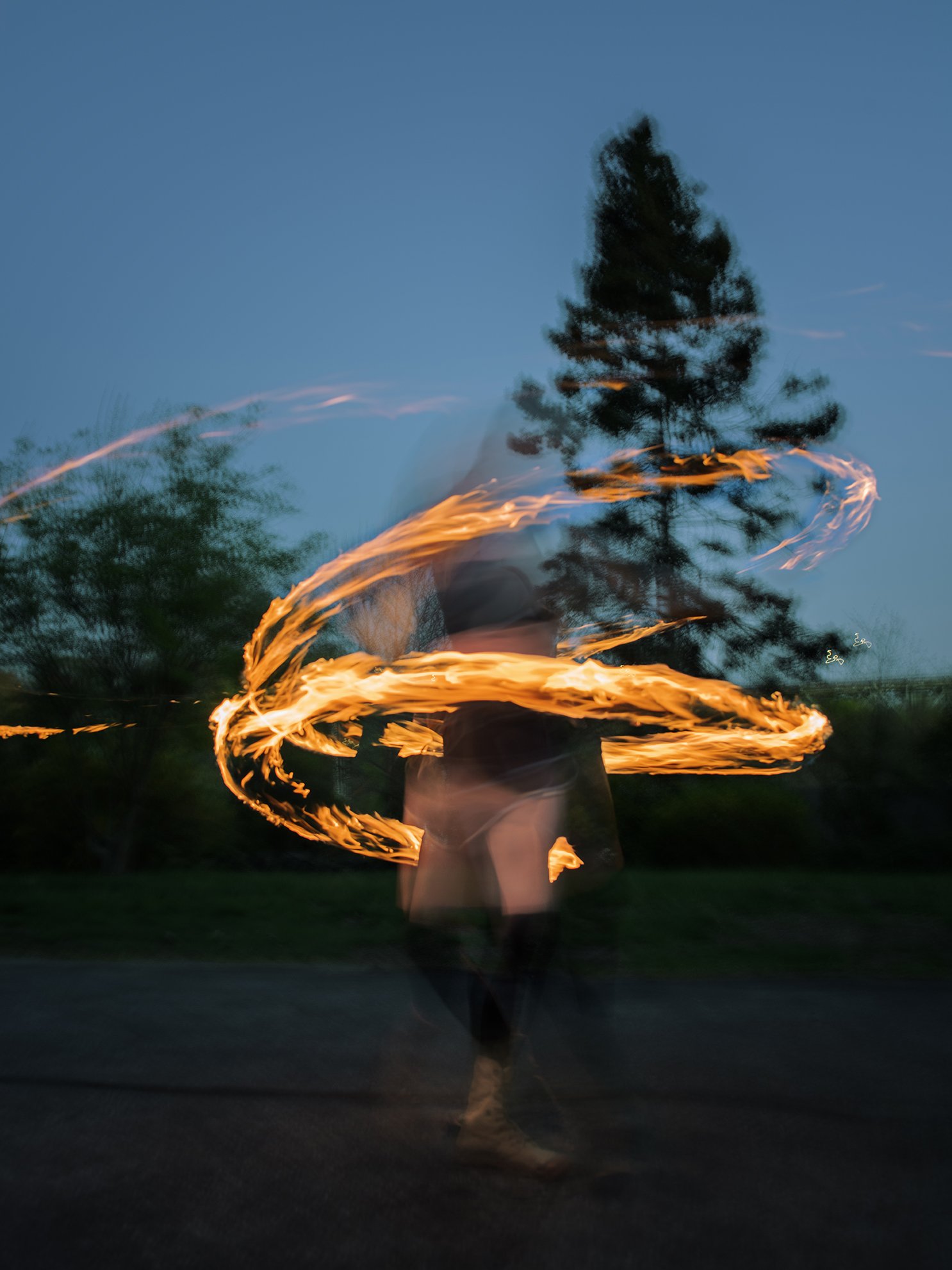 Person performing a fire spinning dance at dusk with a puppy hammer prop, creating flames swirling around them.