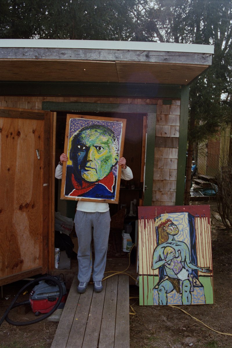 Portrait of an artist holding a colorful painting of a man in front of a shed, with a drawing of a seated woman with a crown on the ground beside them.