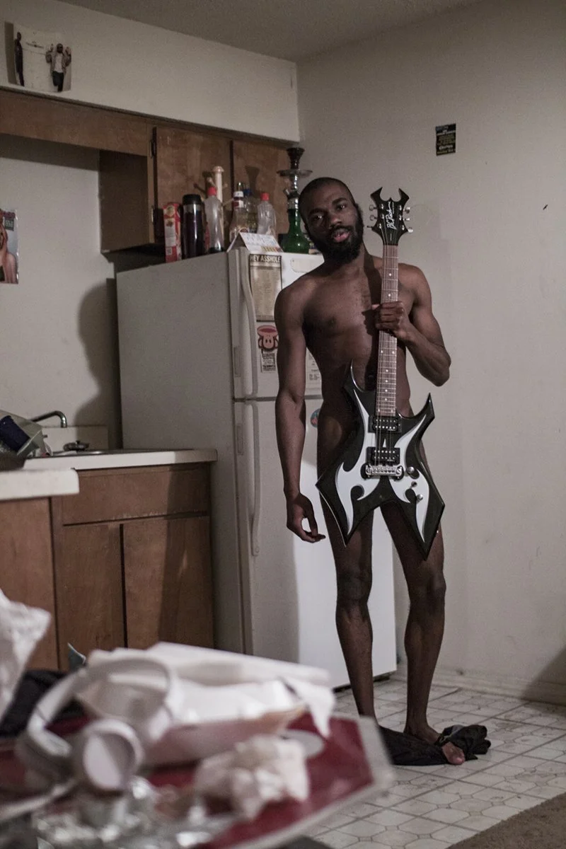 A nude man holding a black and white electric guitar in a kitchen.