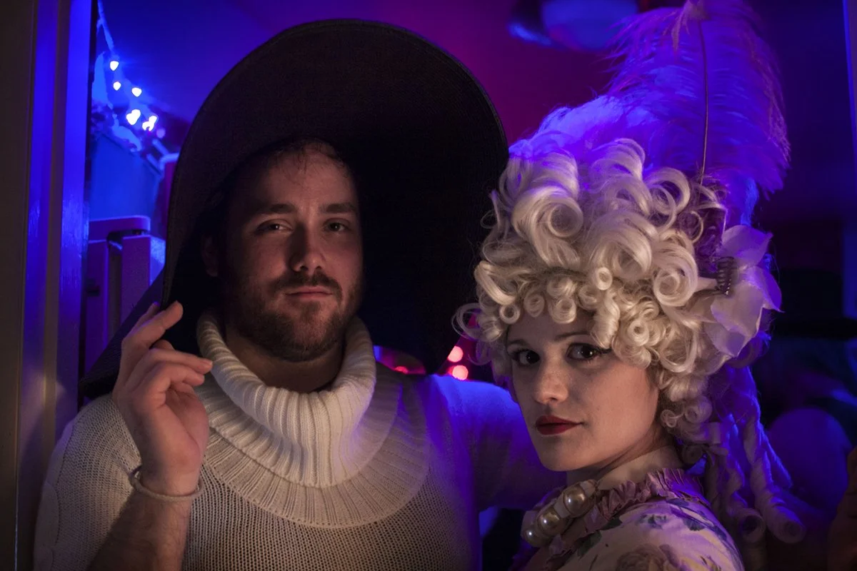 Double portrait from a queer costume party where a man dressed as Audrey Heppburn and a woman dressed as Marie Antoinette pose for a photo with colorful lighting.