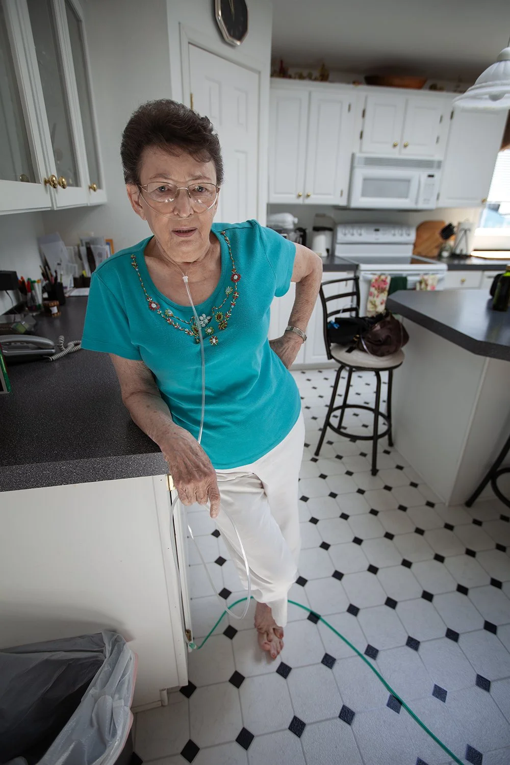 Grandmother with oxygen tube standing in a kitchen, leaning on counter, looking at the camera.