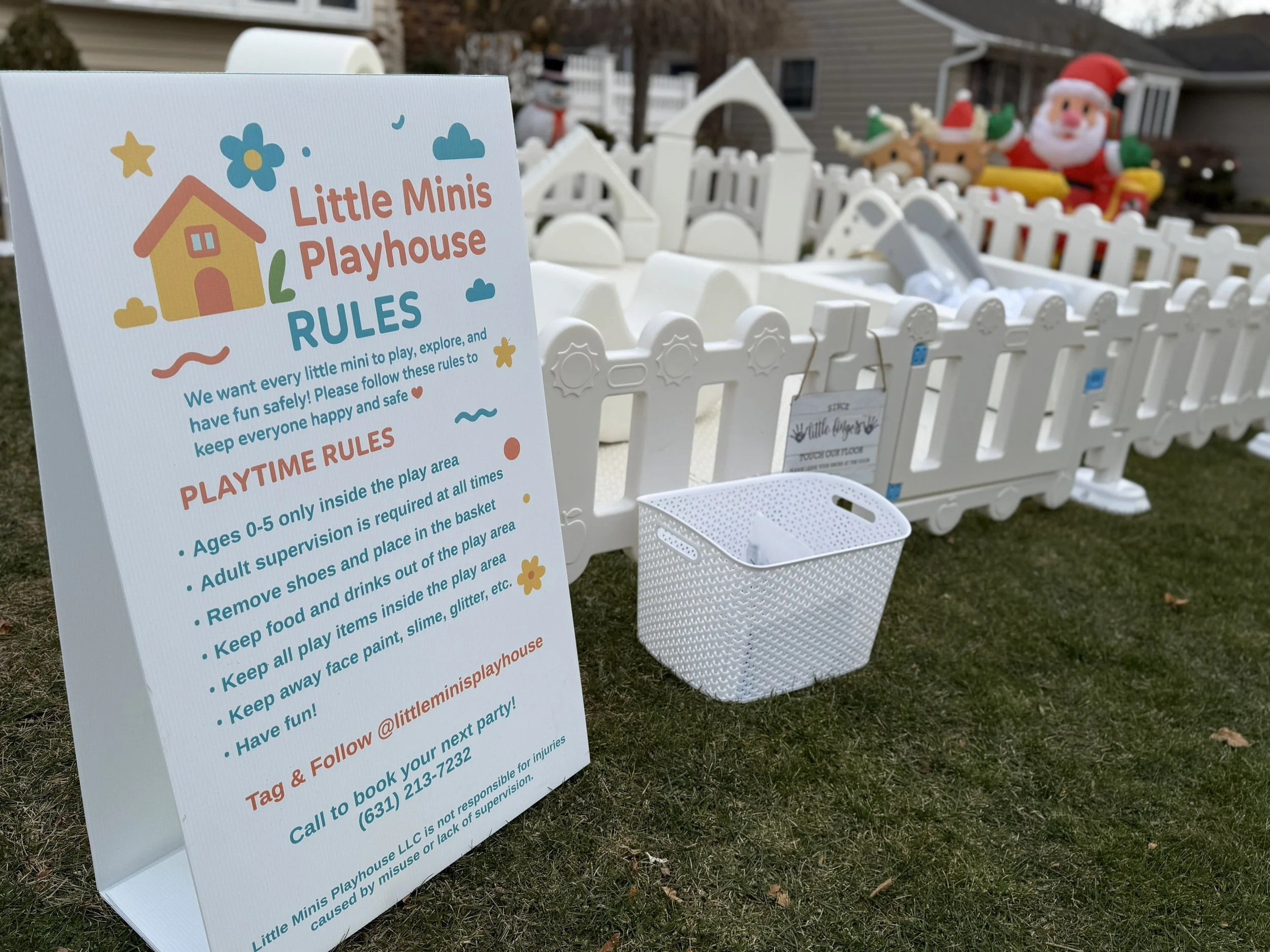A white sign with colorful text and illustrations explaining the rules for Little Minis Playhouse, positioned outside a small fenced play area with Christmas decorations, Santa Claus and reindeer figures in the background.