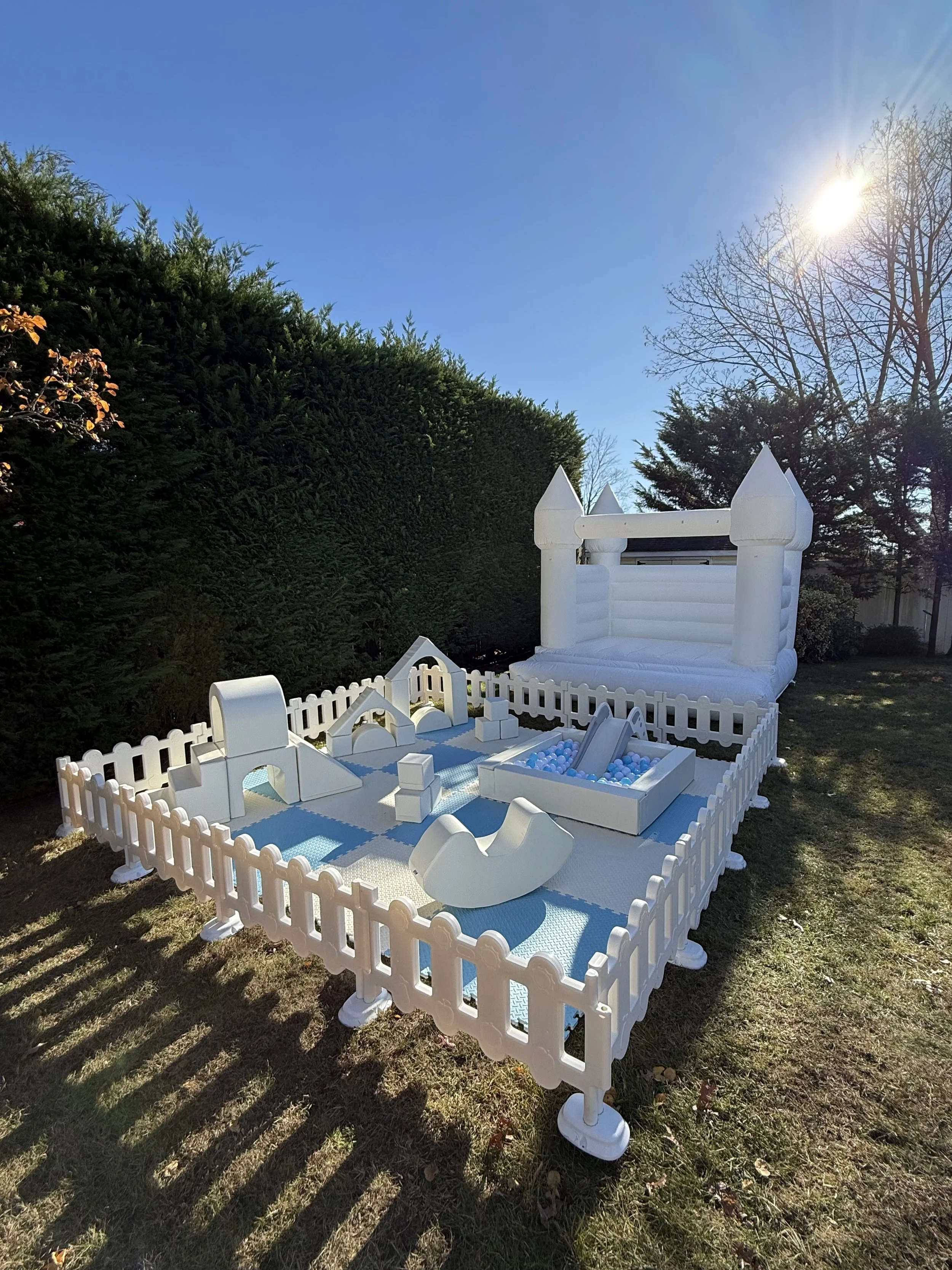 White inflatable bounce house with a castle design, including towers and a slide, set up in a backyard with a play area that has a sandbox filled with blue and white balls, surrounded by a white picket fence. Trees are visible in the background under a clear blue sky with the sun shining.
