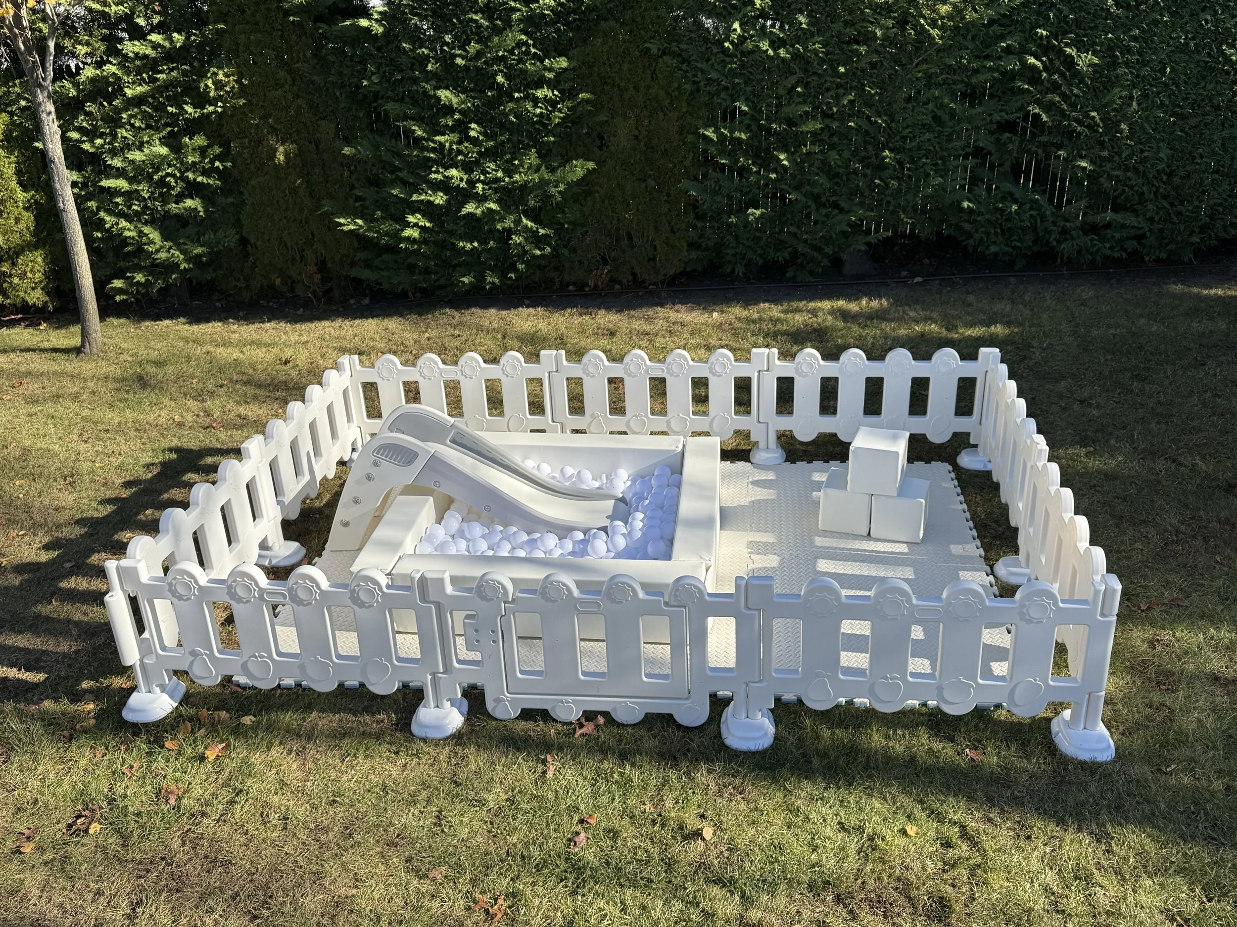 A small white kids' play area with a mini slide, balls, and soft blocks, surrounded by a white fence on a grassy lawn with trees in the background.