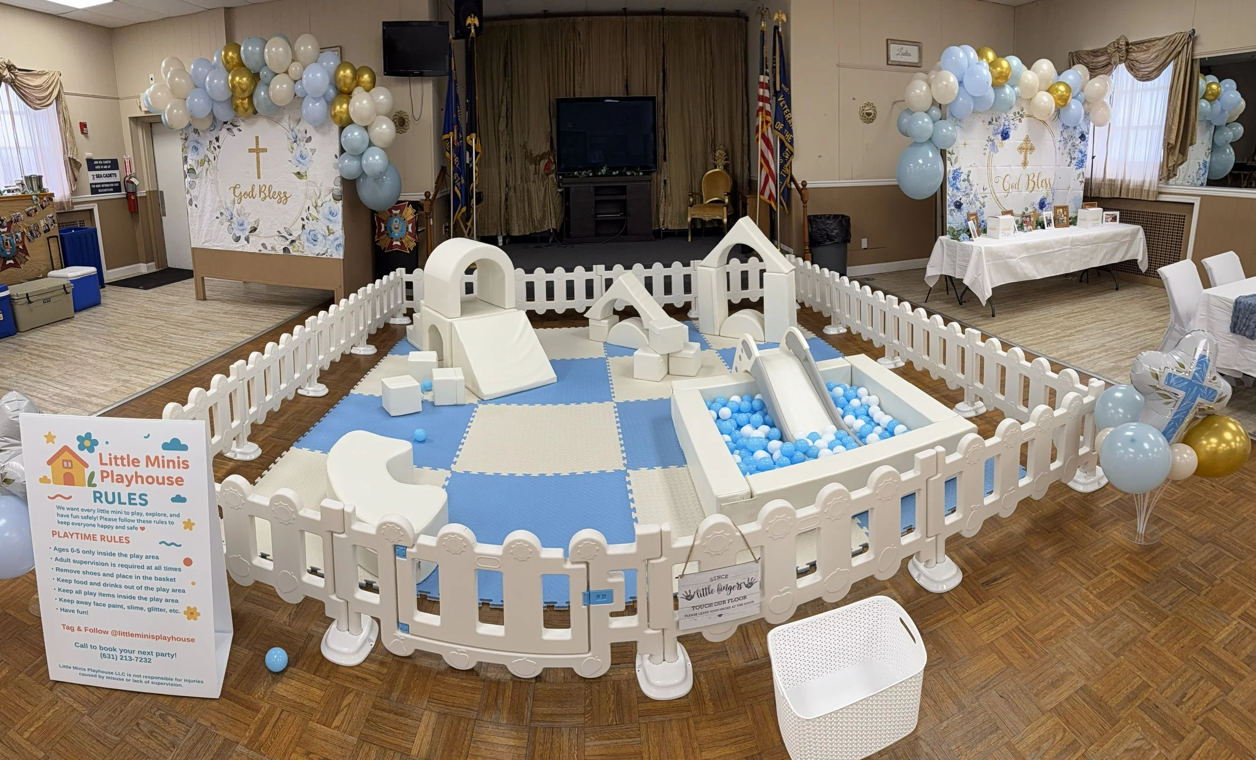 Indoor play area for children with a white picket fence, foam blocks, slides, and a ball pit filled with blue and white balls, decorated with balloons and religious-themed banners.