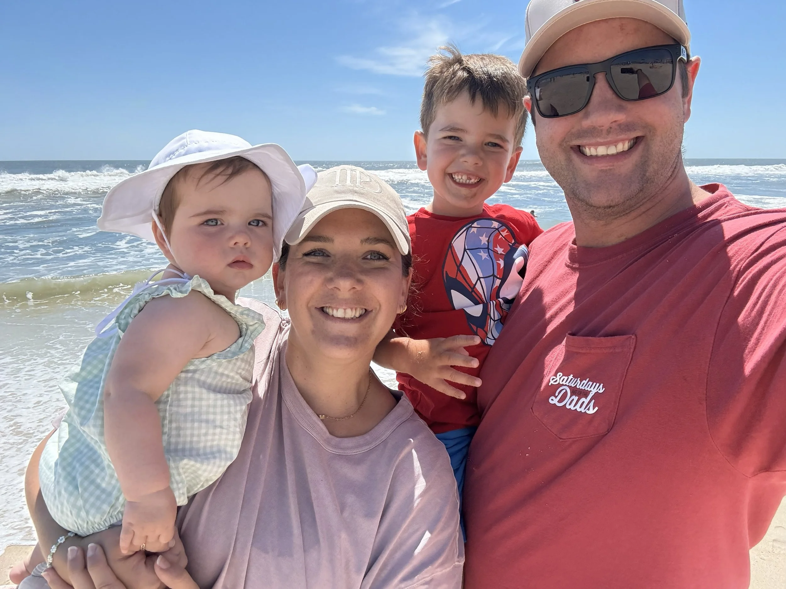 A family of four at the beach taking a selfie, with the ocean and waves in the background. The father is wearing sunglasses and a red shirt that says 'Saturdays Dads.' The mother is smiling and wearing a light-colored shirt and a white cap. The little girl is on the mother's arm, wearing a white floppy hat and a light outfit, with a serious expression. The young boy is next to the father, smiling, wearing a red shirt with a graphic design.