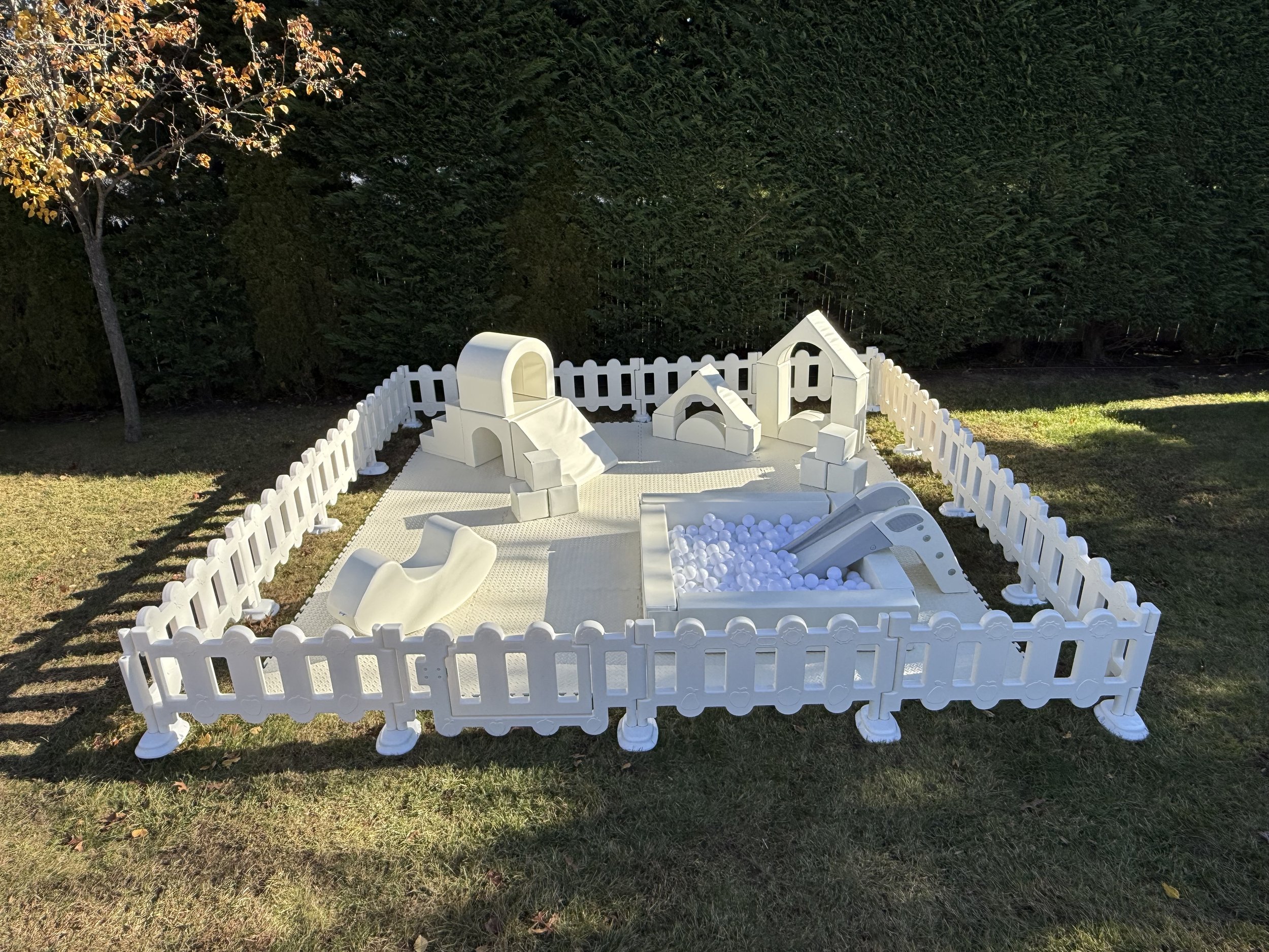 White outdoor playground with slides, ball pit, and small houses, surrounded by a white picket fence on grass and trees.