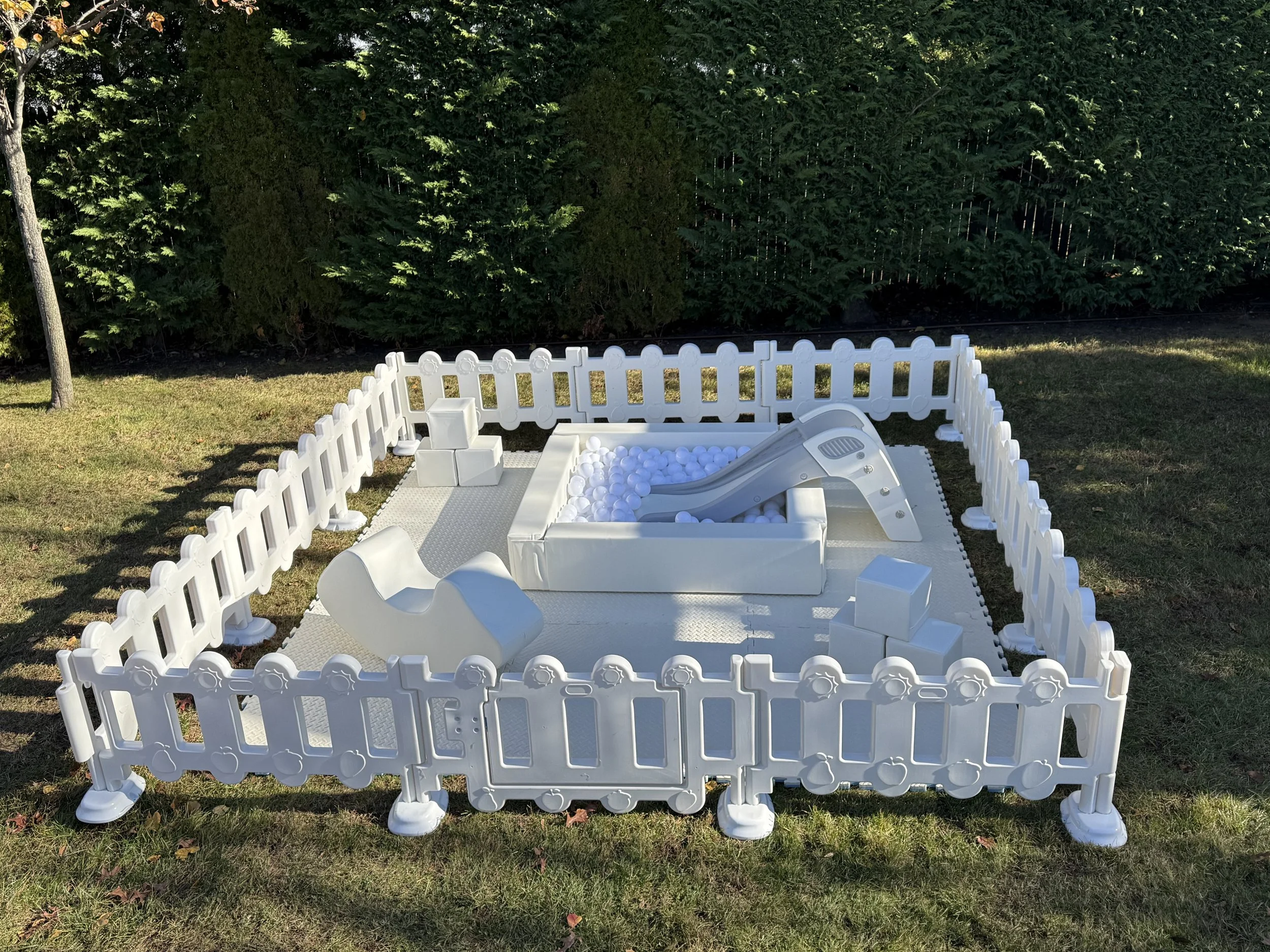 A white plastic play area for children with a small slide, ball pit filled with white balls, and various blocks and toys, surrounded by a white picket fence on a grassy lawn with trees in the background.