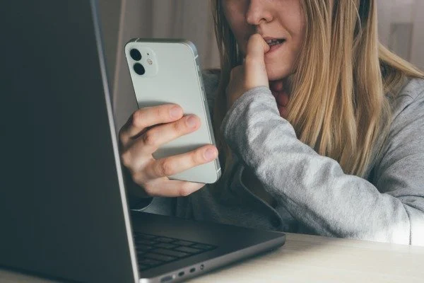 Candid image of a blonde woman biting her thumb while reading something on her smartphone, with a laptop in front of her. Depicts anxiety, stress.