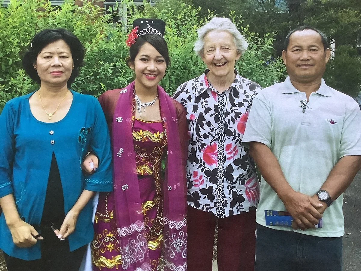 Four women and one man standing outdoors in front of green foliage, smiling for the camera. The woman second from the left wears traditional attire and a tiara, suggesting a ceremonial or cultural event.