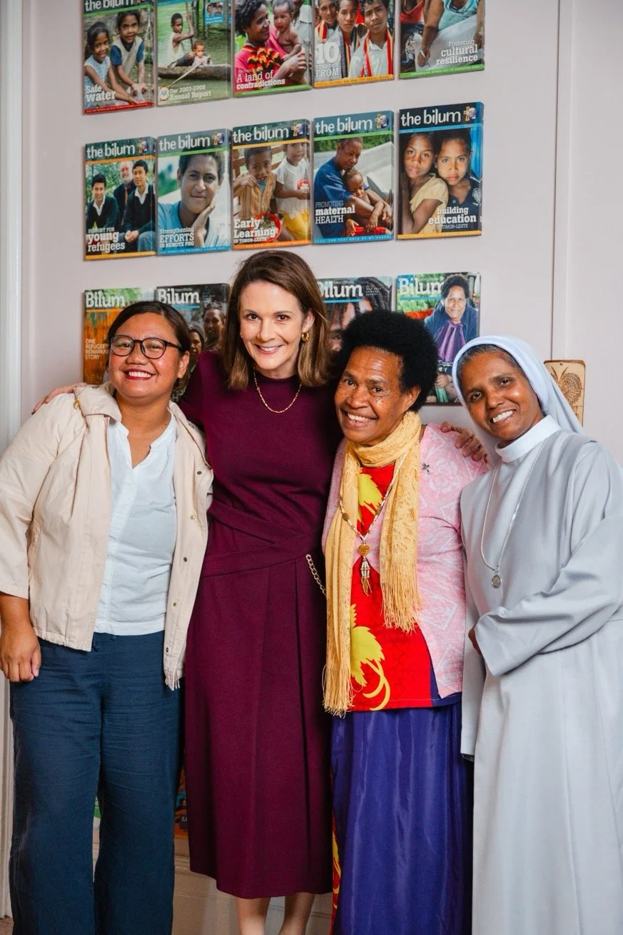 Four women of diverse backgrounds smile together in front of a wall displaying multiple issues of The Bilum, a publication highlighting stories of cultural resilience, maternal health, early education, and refugee support in the Pacific region.