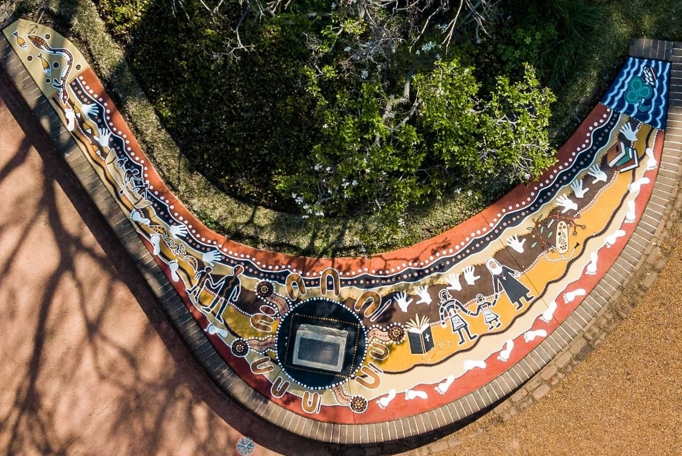 Aerial view of a vibrant Aboriginal mural painted on a curved concrete path, featuring traditional dot painting, white handprints, and symbolic figures representing community, education, and cultural storytelling