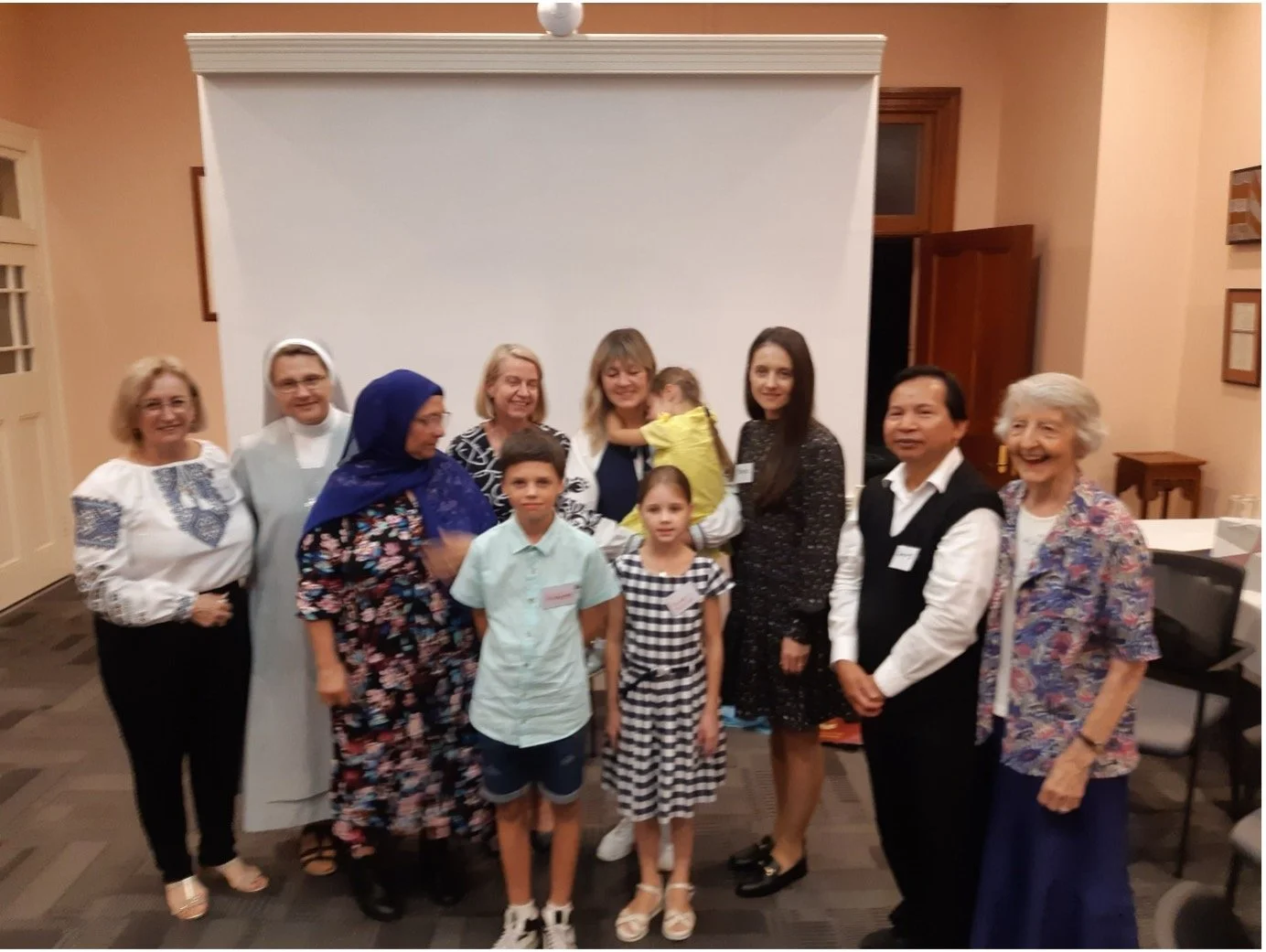 A group of people gathered in a room with a white backdrop, including children, women, and a nun, some smiling and posing for the photo.