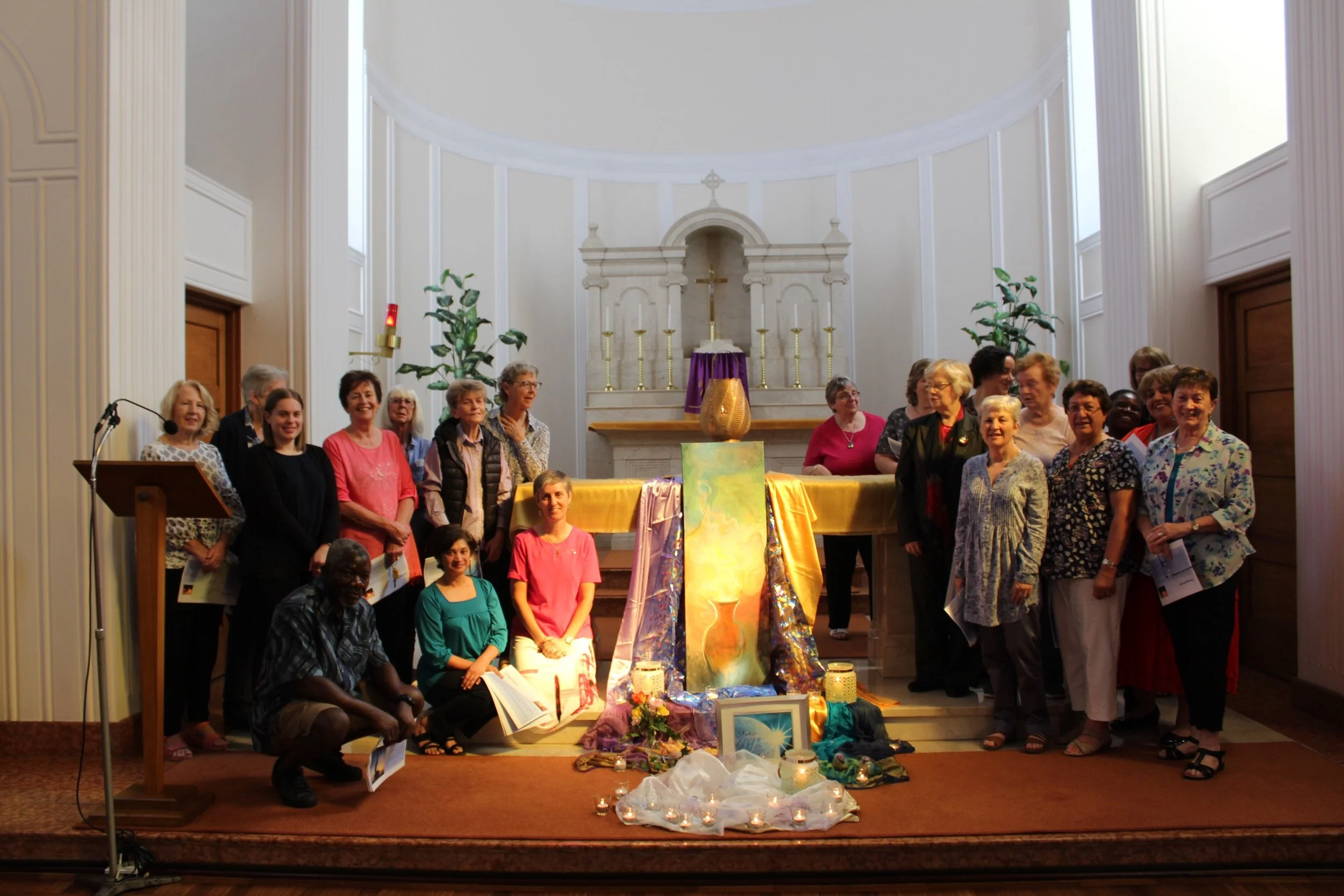 Group of people gathered in a church for an event, standing in front of a decorated altar with candles and art pieces.