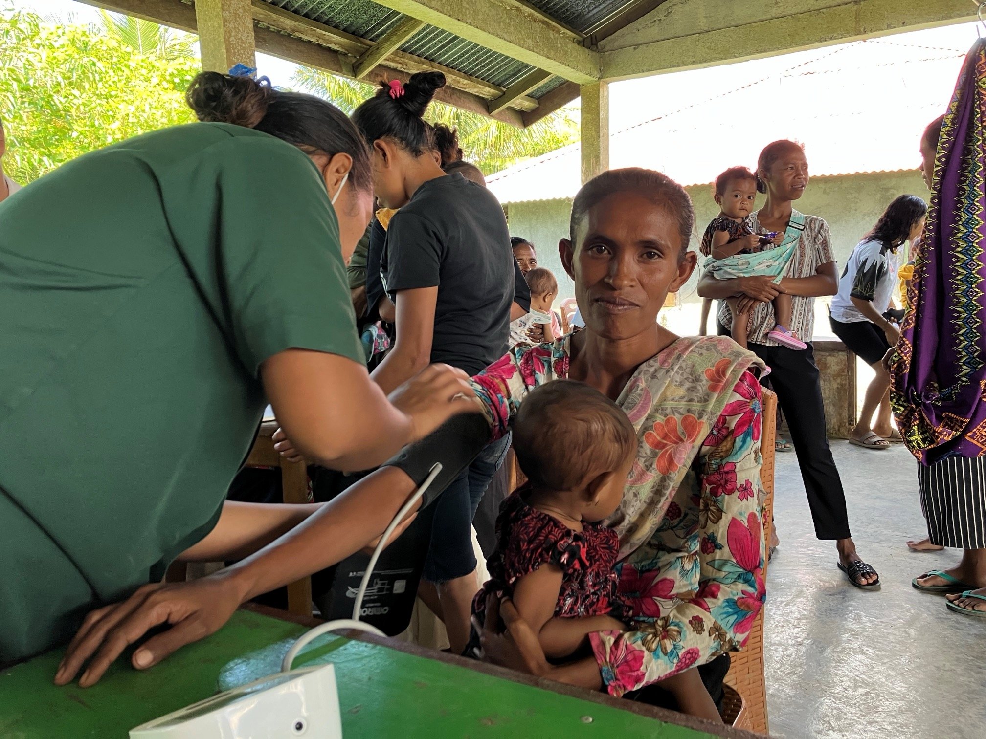 A healthcare worker in a green uniform takes the blood pressure of a woman holding a baby in her lap at a community clinic in a tropical setting.