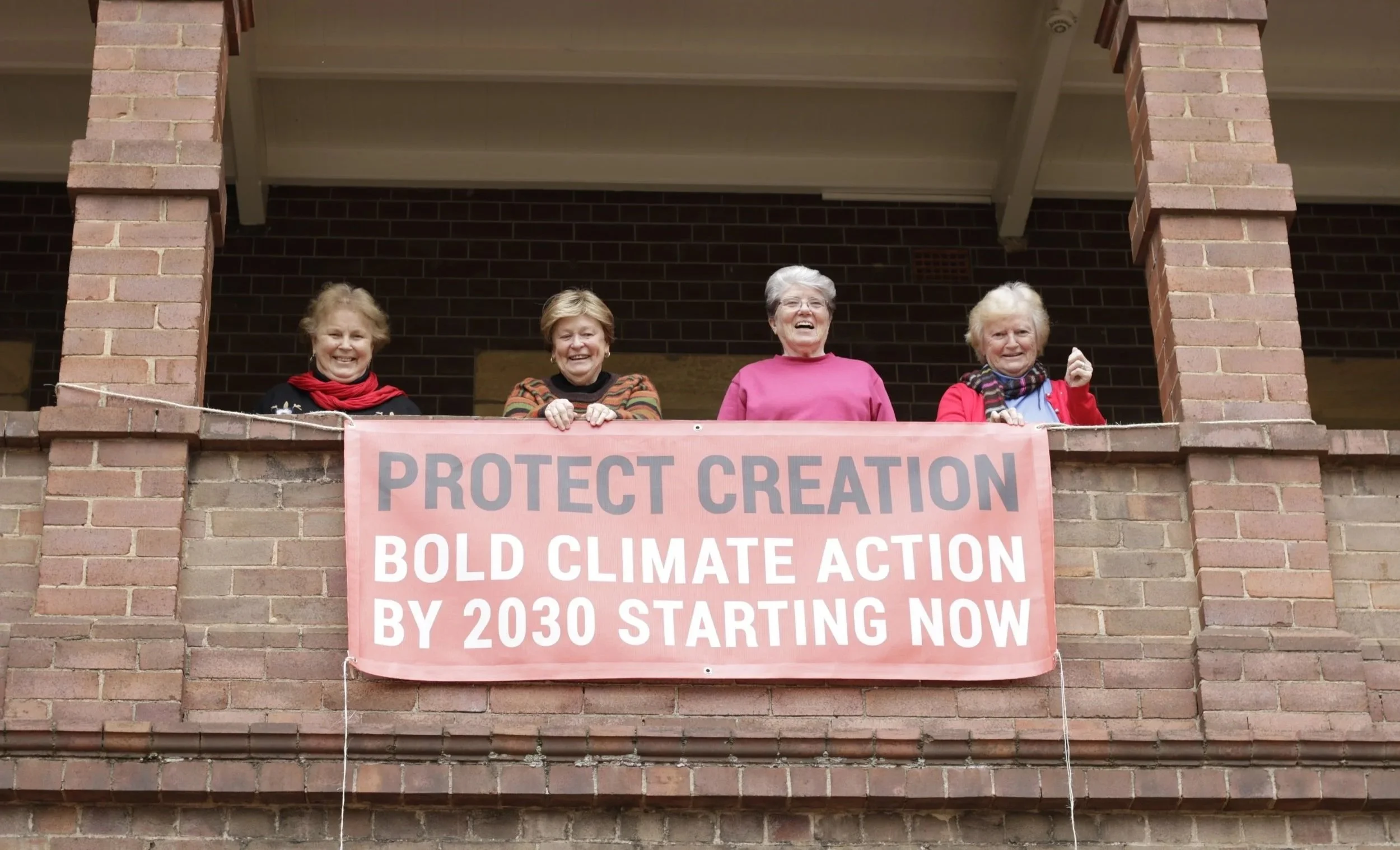 Four elderly women standing on a brick balcony holding a pink banner that reads "Protect Creation, Bold Climate Action by 2030 Starting Now"