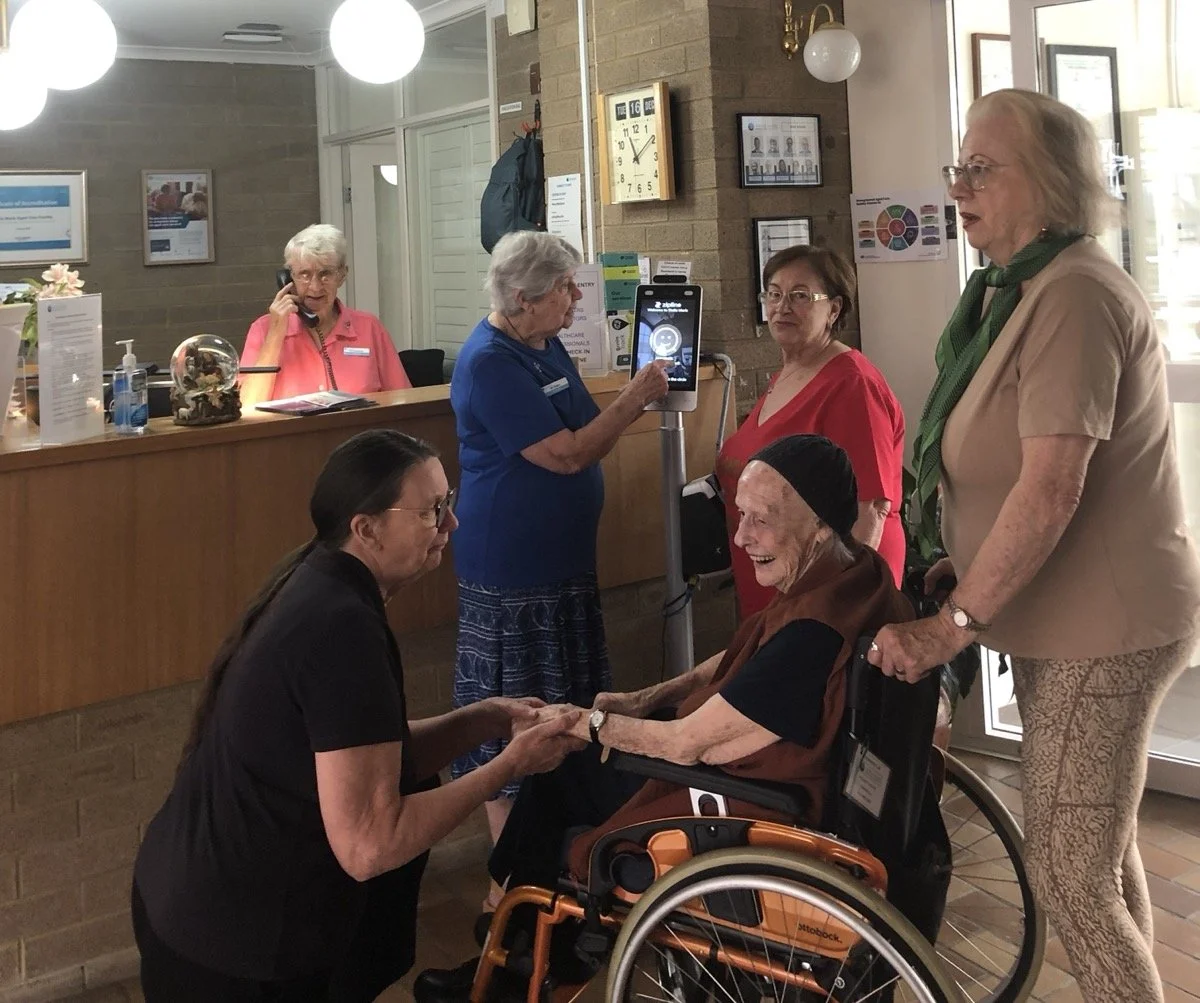 A group of older women interacting in a lobby or reception area, one woman in a wheelchair holding hands with a woman kneeling, in front of a reception desk with a staff member talking on the phone.