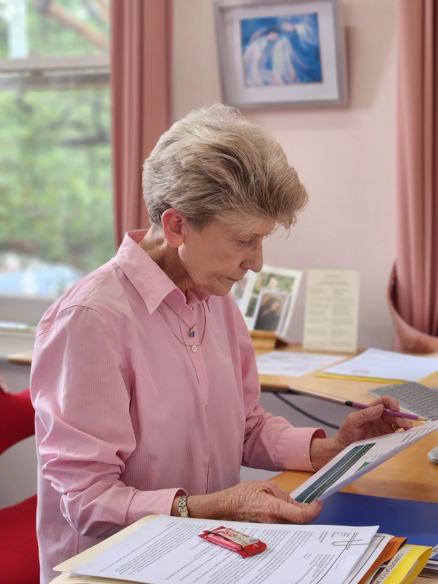 An elderly woman with short gray hair, wearing a pink button-up shirt, is sitting at a desk, reading a document with papers, pens, and other items on the desk. There are pink curtains and a framed picture of ocean waves hanging on the wall behind her.