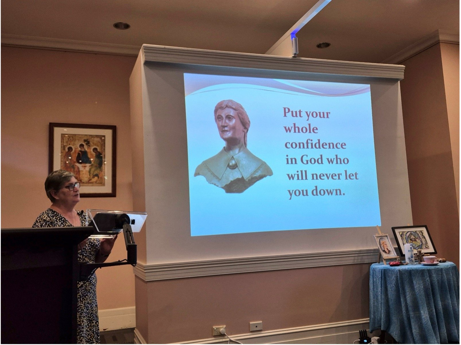 A woman gives a talk at a podium beside a large projection of a quote that reads, “Put your whole confidence in God who will never let you down.”