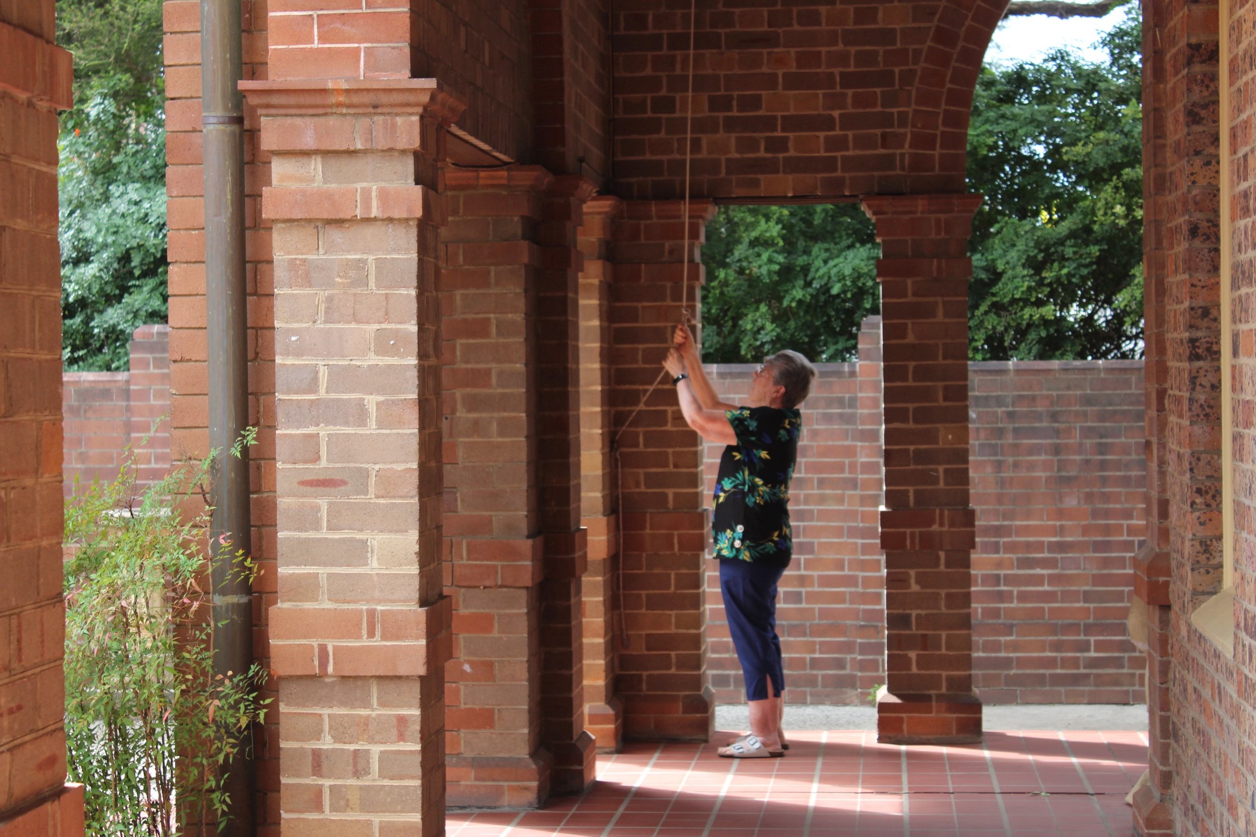 An elderly man wearing a black floral shirt and navy pants pulling a bell string in a brick colonnade.