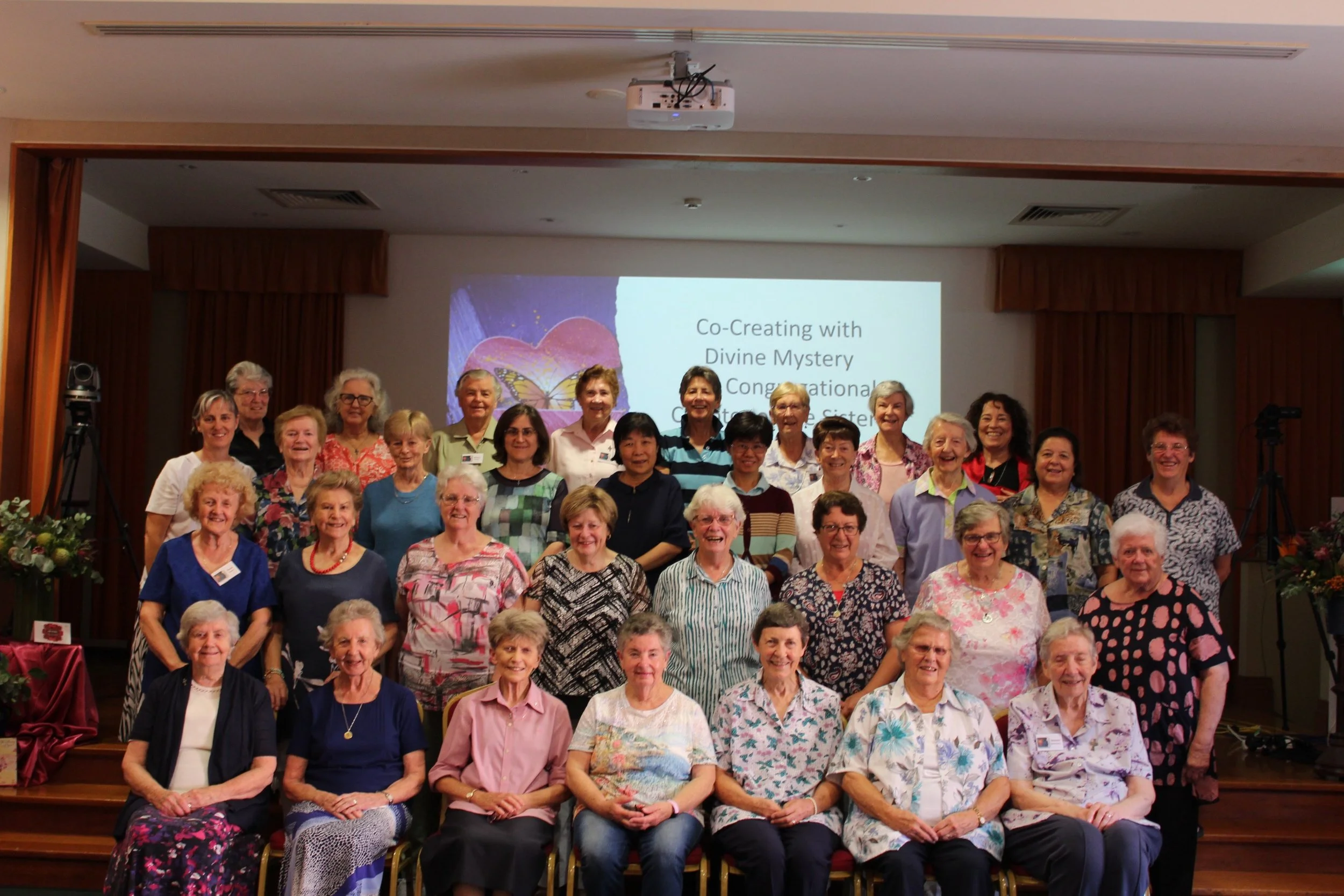 Group of women and men gathered on stage for a celebration or photo, with a presentation slide behind them that reads, 'Co-Creating with Divine Mystery Congratulations,' in a conference or event room.