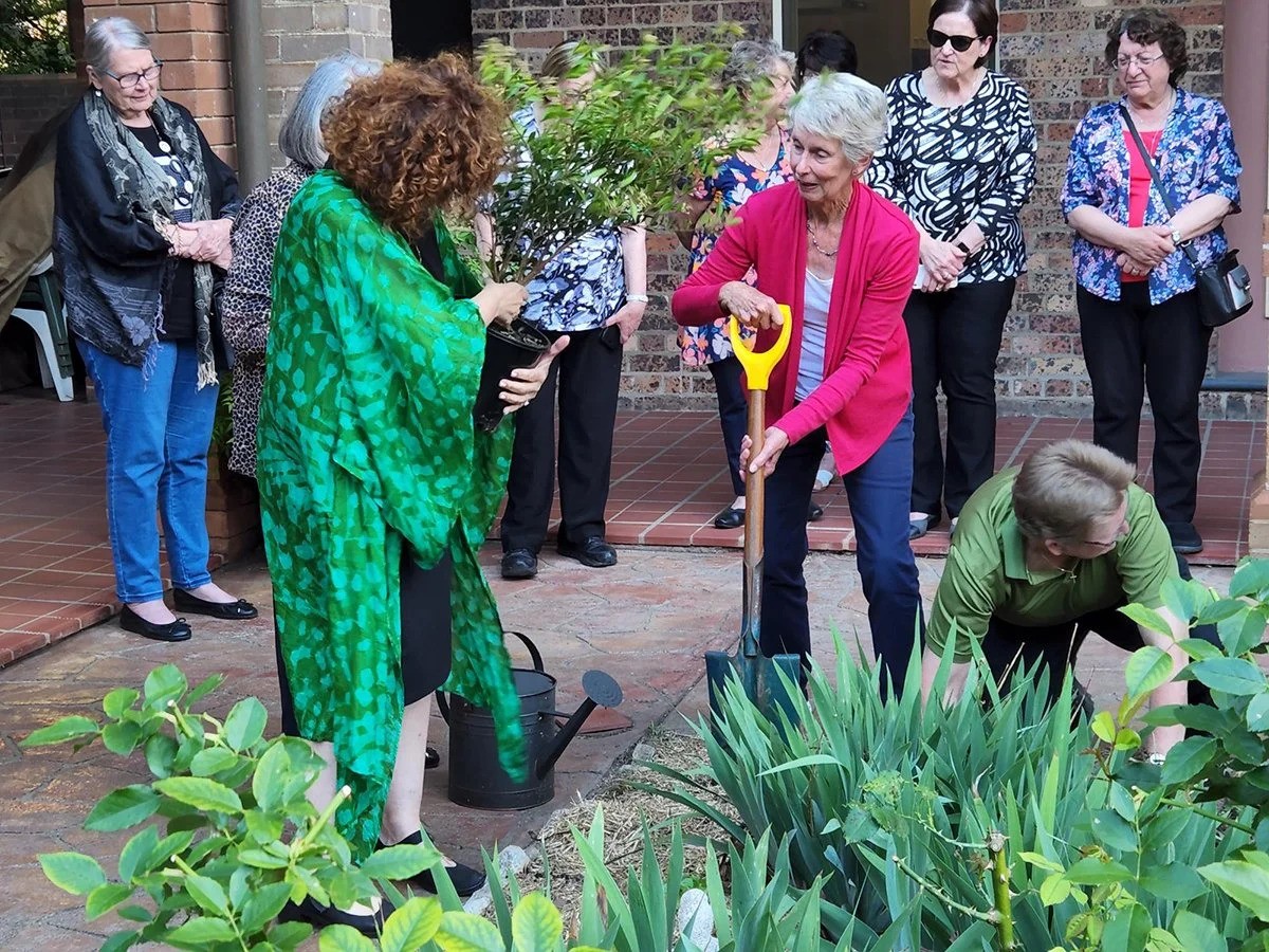 Group of women participating in a ceremonial tree planting event in a garden courtyard. A woman in a bright pink cardigan is holding a yellow-handled shovel, assisted by another woman in green.