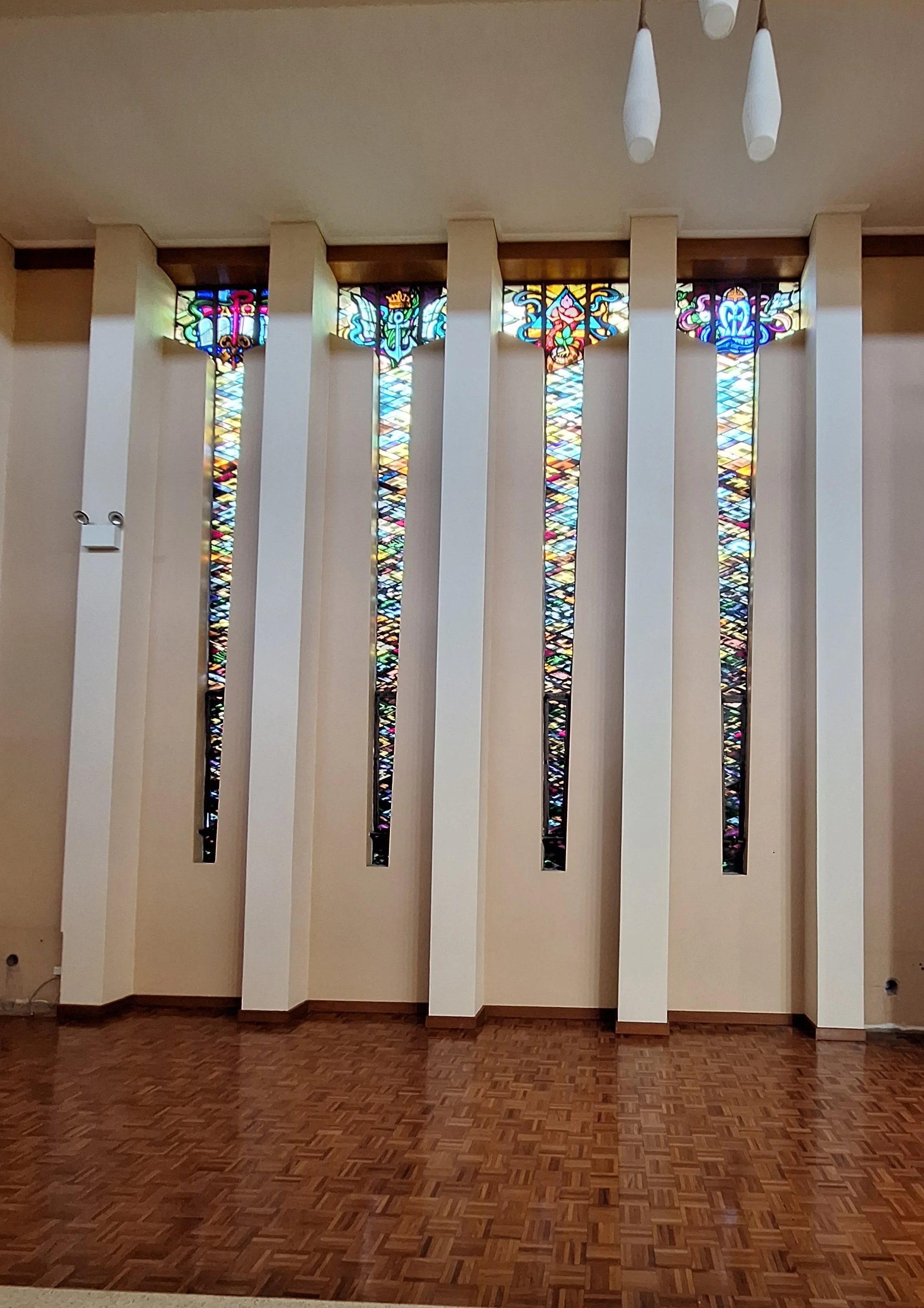 Colorful stained glass windows with intricate designs, located in a modern interior with white vertical panels and a wooden parquet floor.