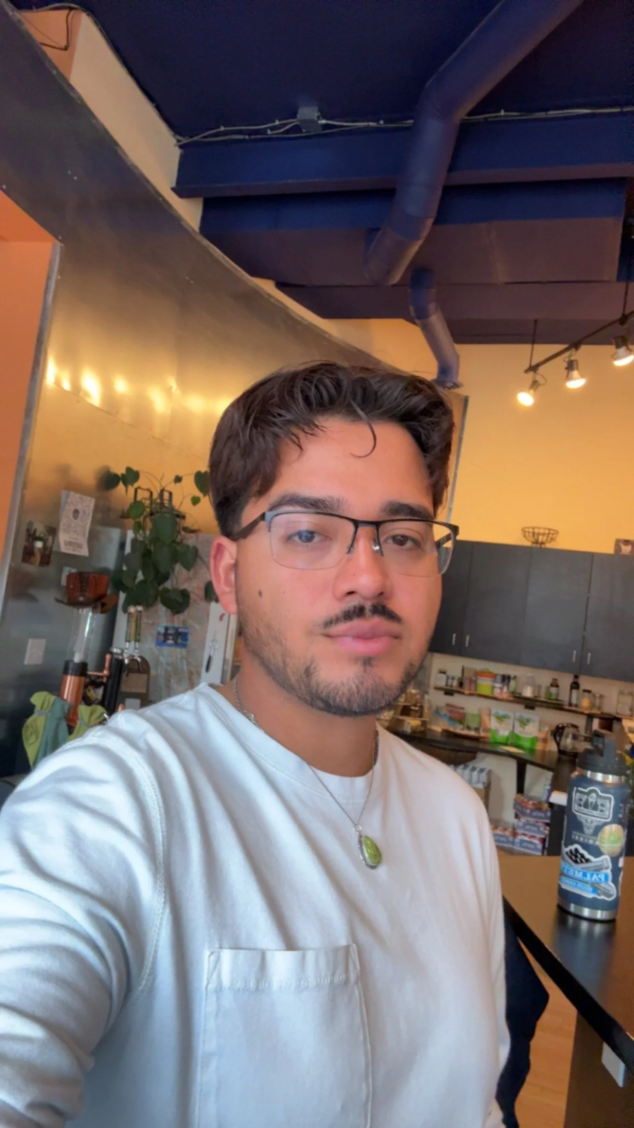 A young man with glasses, facial hair, and a green pendant necklace taking a selfie indoors, with a kitchen and a coffee station in the background, and warm lighting.