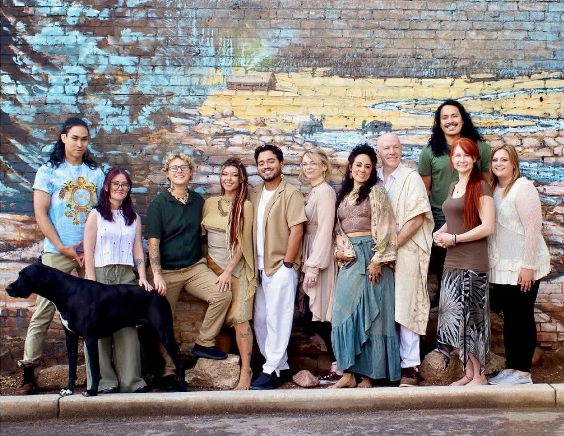 A diverse group of people, including men and women of various ages and ethnicities, posing outdoors in front of a colorful mural on a brick wall. They are smiling and standing close together.