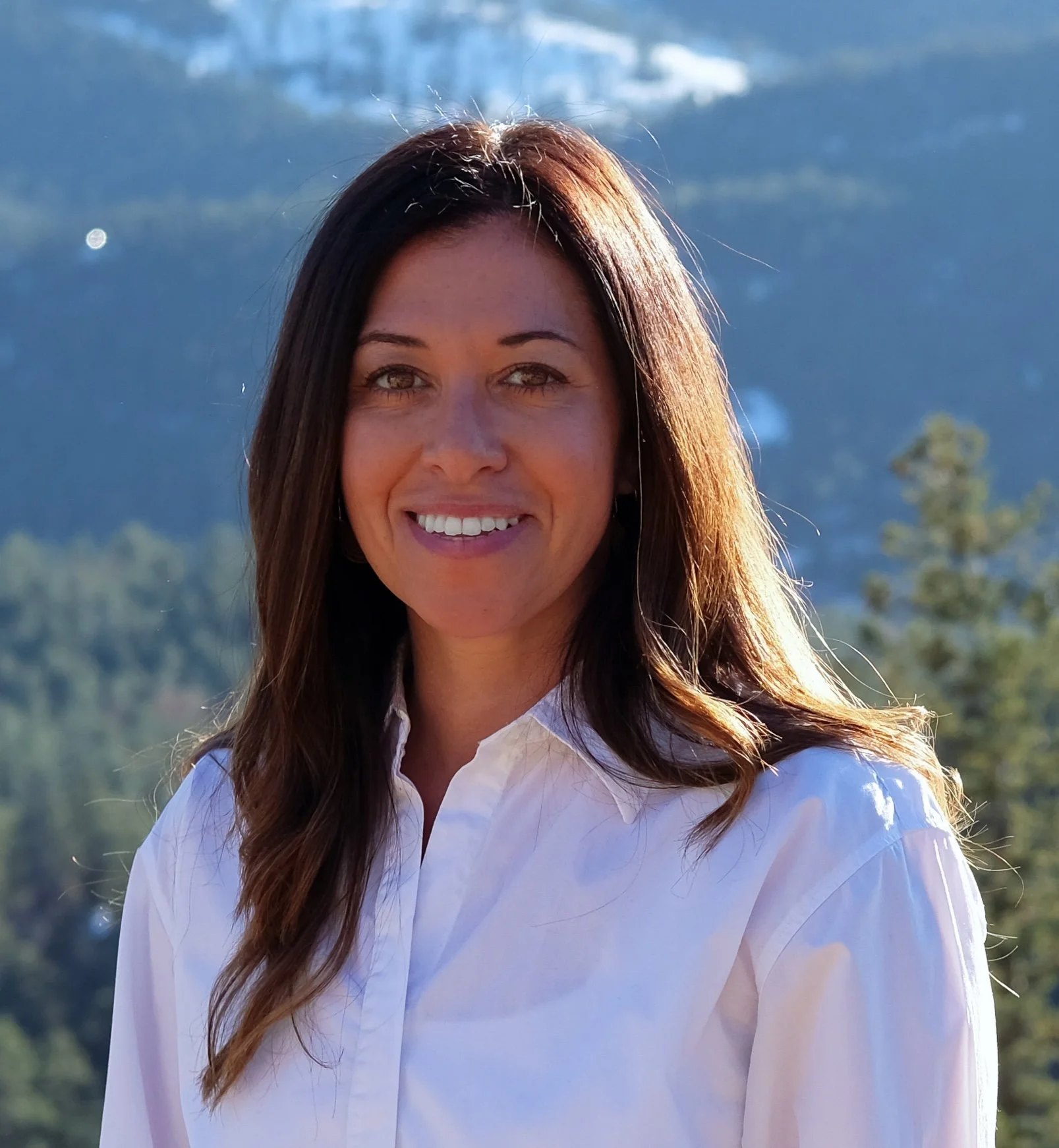 A woman with long brown hair wearing a white shirt smiling outdoors with mountains and trees in the background.