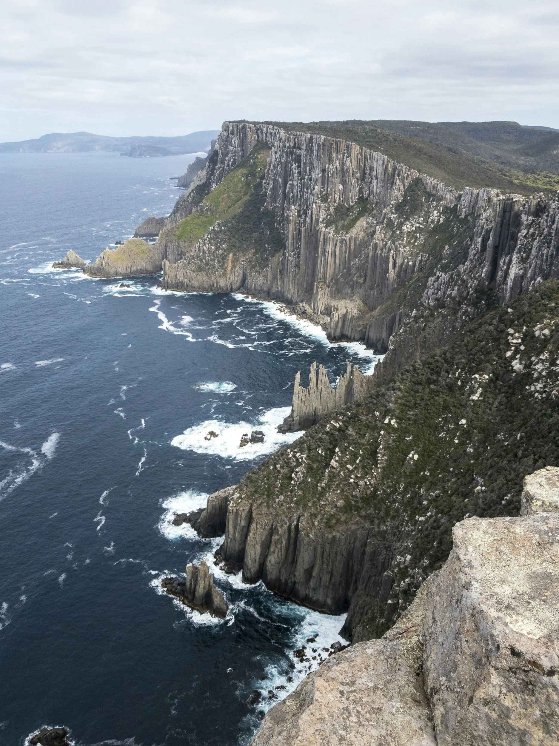 Cliffs along a rugged coastline with rocky outcroppings and waves crashing at the base, under a cloudy sky.